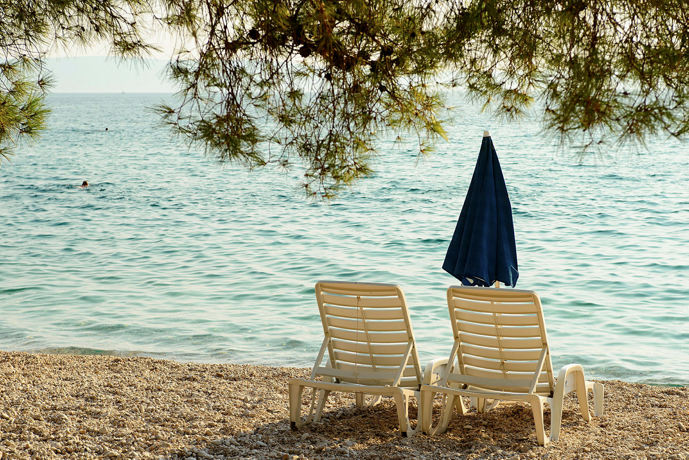 To solsenger og en parasoll ved en steinete strand med sjøen i bakgrunnen. (AI-generert tekst)