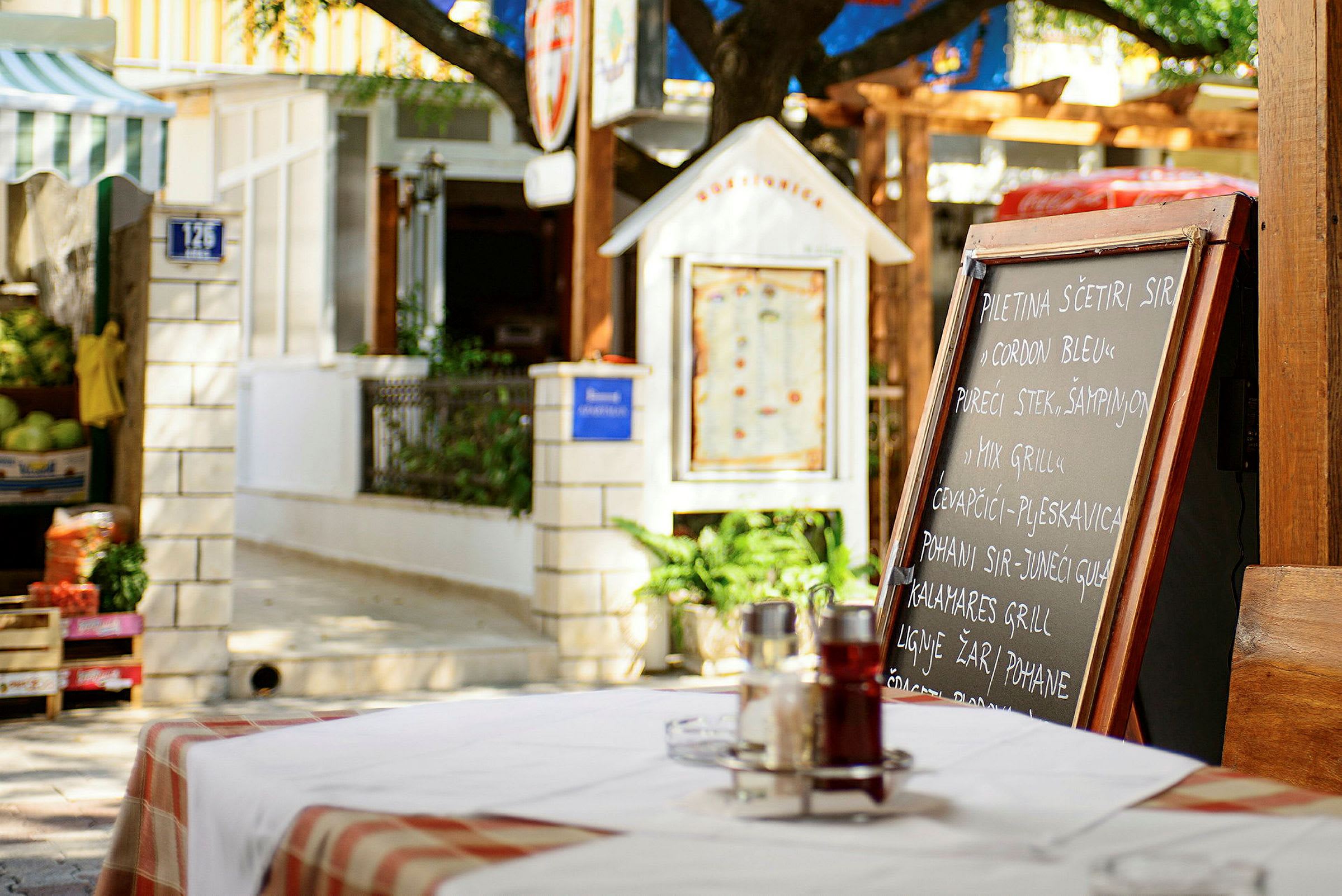 En restaurantmeny på en svart tavle utendørs på et bord med en rødhvit duk, omgitt av grønt og bygninger. (AI-generert tekst)