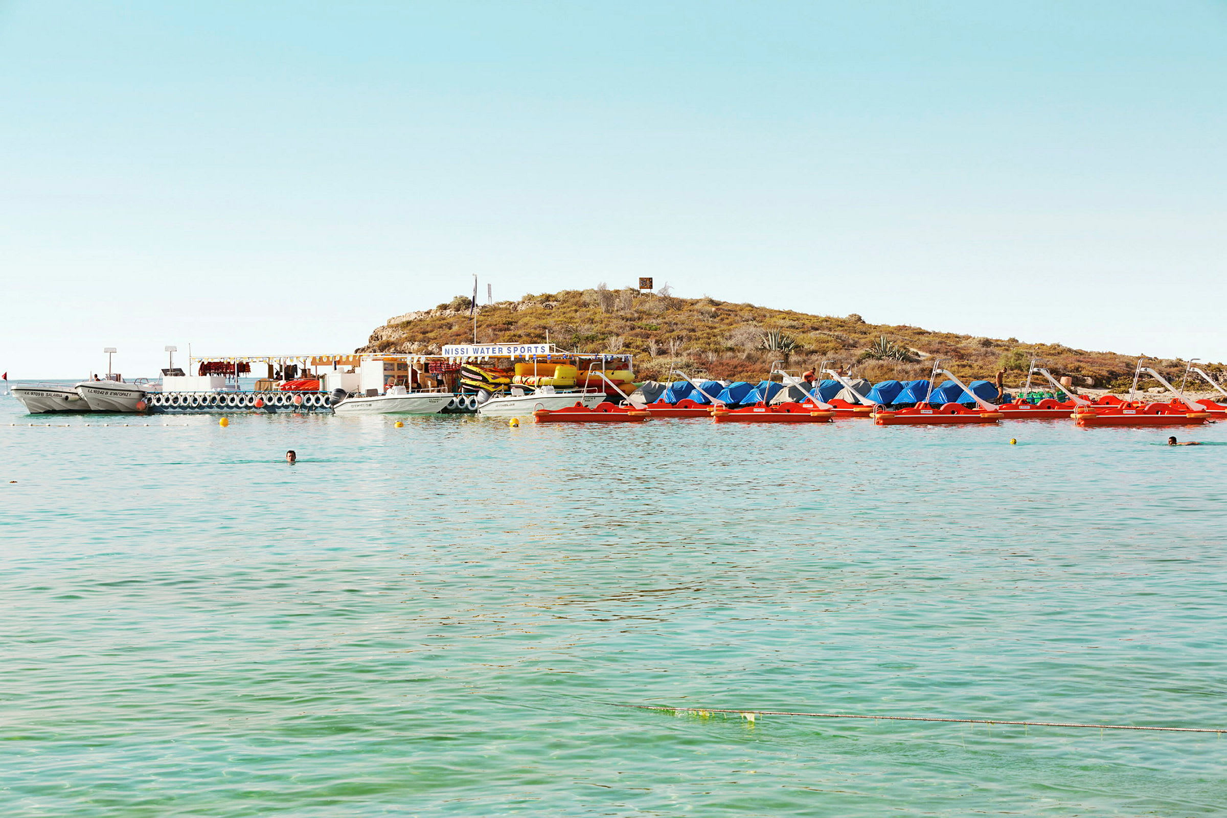 En samling av båter og tråkkebåter ved en strand med grønt vann og en liten høyde i bakgrunnen. (AI-generert tekst)