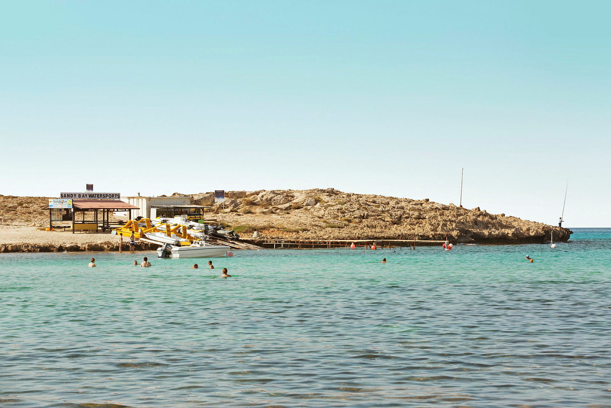 En liten bukt med klart vann. Folk bader, og båter ligger fortøyd ved en brygge. En vannsportanlegg med utstyr ligger på stranden. (AI-generert tekst)