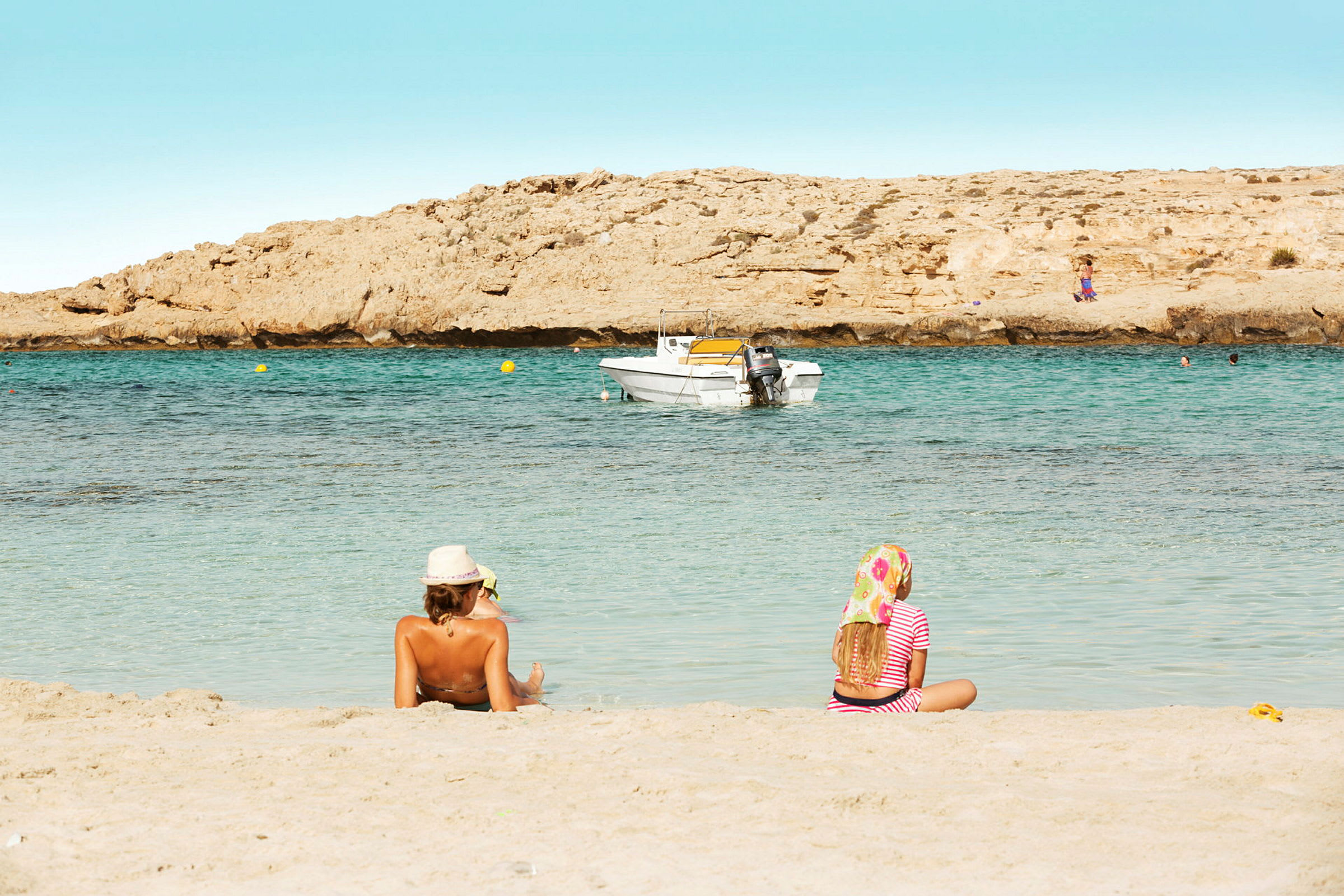 To personer sitter på en sandstrand med havet og en båt i bakgrunnen. (AI-generert tekst)