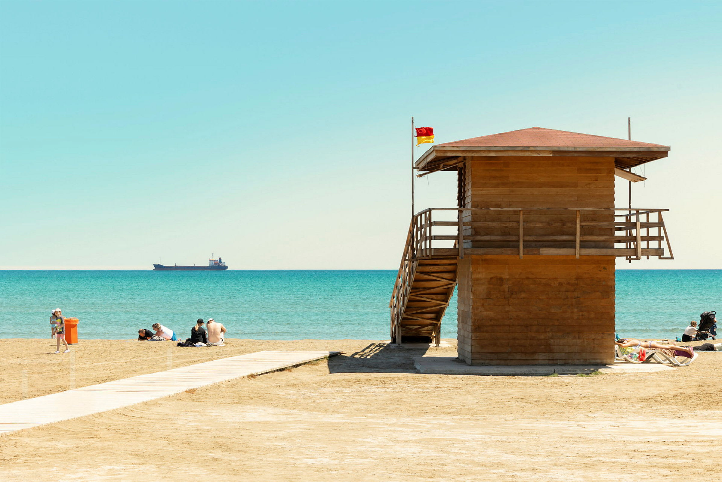 En strand med et livreddertårn, noen mennesker som sitter ved vannkanten og et skip på havet. (AI-generert tekst)