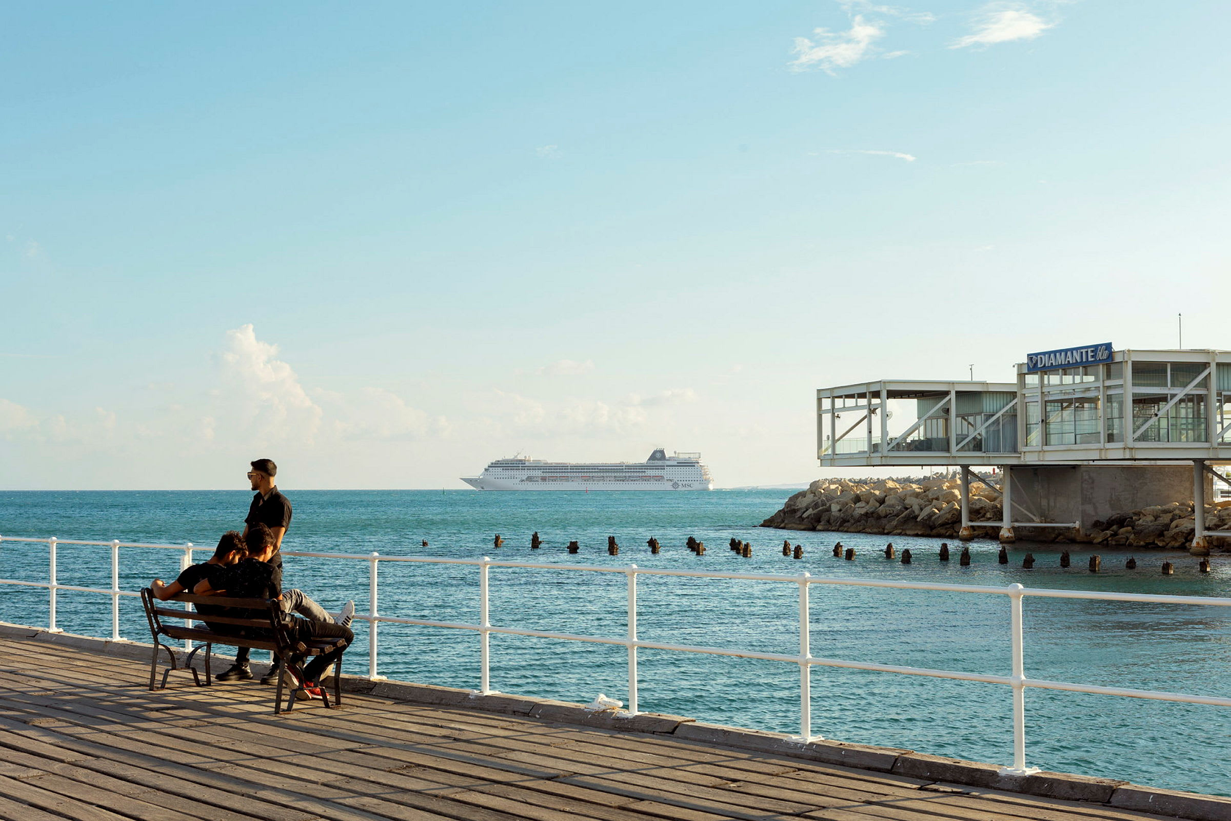 En strandpromenade med en benk, havet og et cruiseskip på horisonten. (AI-generert tekst)