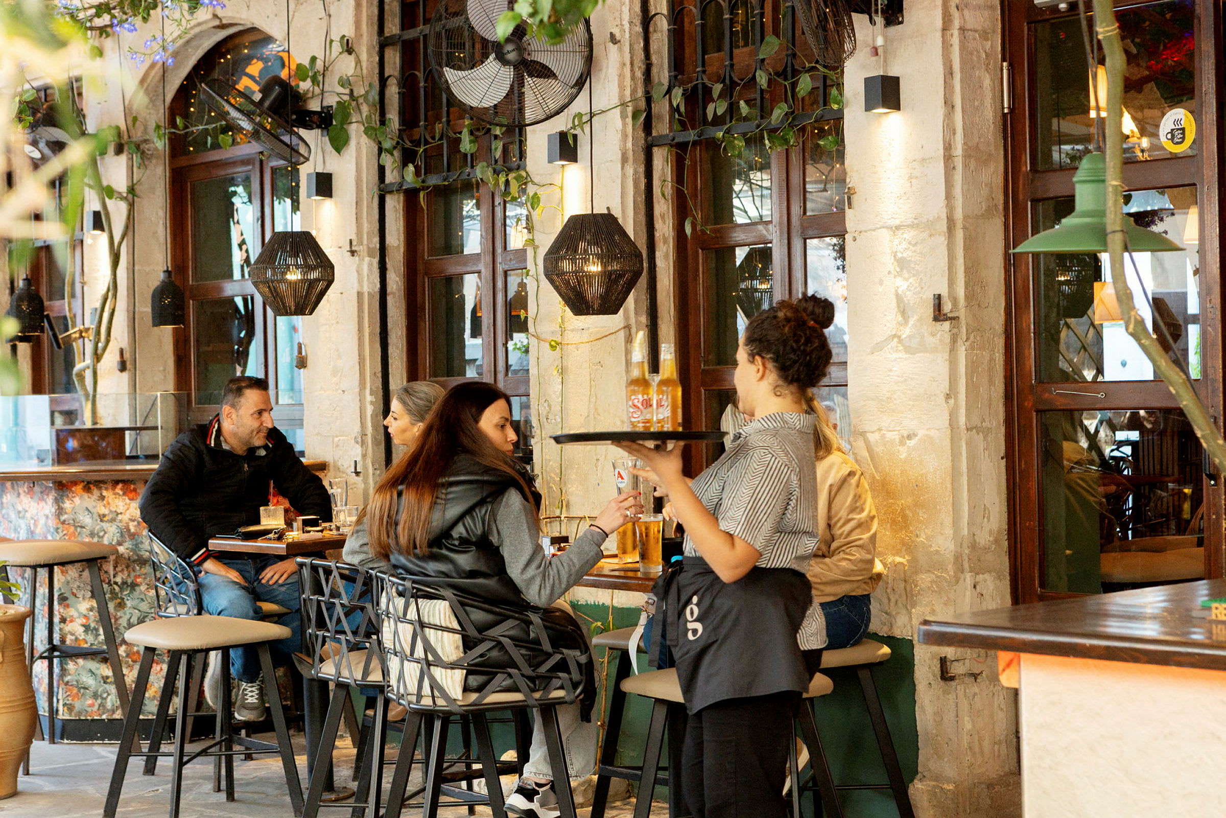 En servitør serverer drikke til gjester i en restaurant med murvegger og vinduer. (AI-generert tekst)