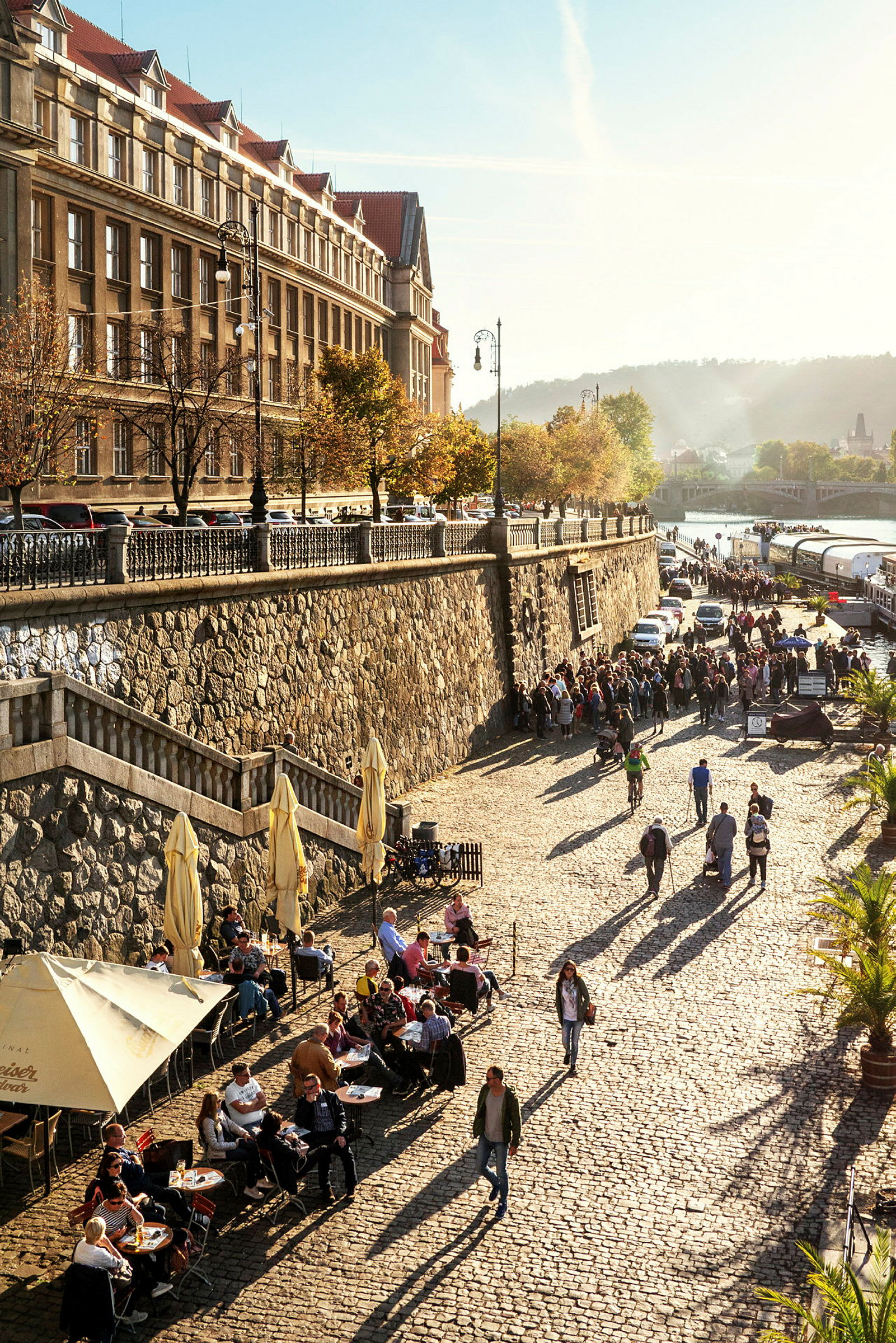 Gatescene med brosteinbelagt vei ved en vannmasse, omgitt av en steinmur og bygninger, med folk som går og sitter ved bord. (AI-generert tekst)
