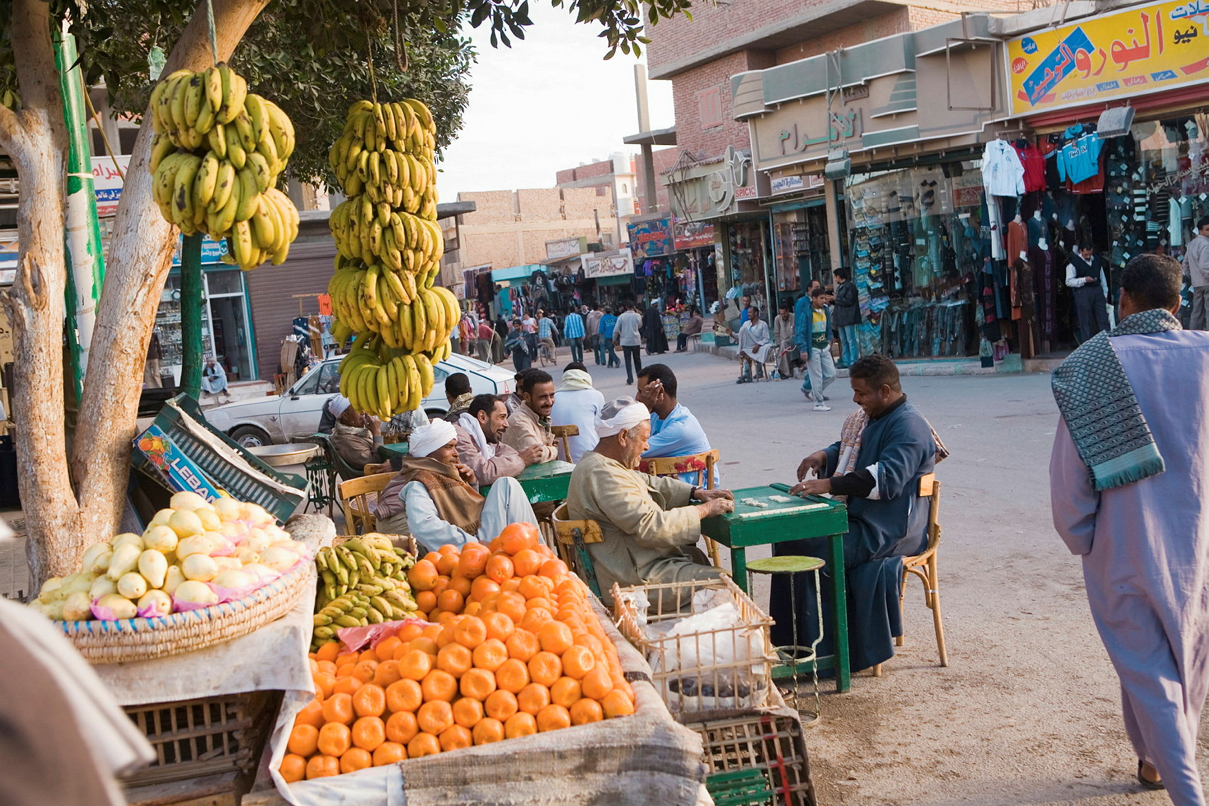 En livlig gate med salgsboder som tilbyr fersk frukt, som appelsiner og bananer. Folk sitter ved bord og prater i en avslappet atmosfære. (AI-generert tekst)