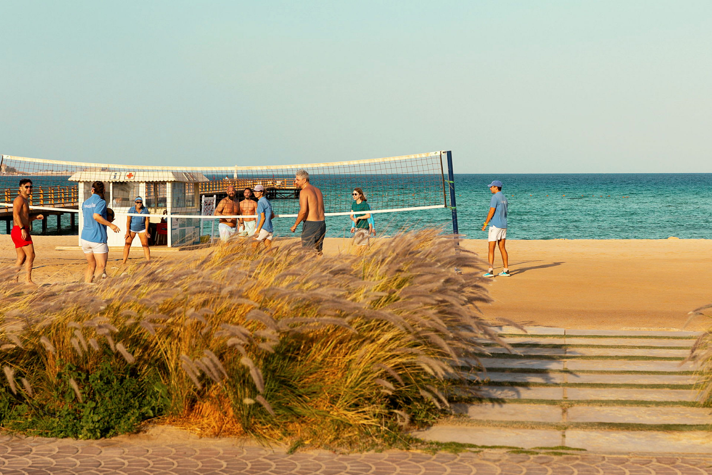 Personer spiller strandvolleyball ved havet, med gress i forgrunnen. (AI-generert tekst)