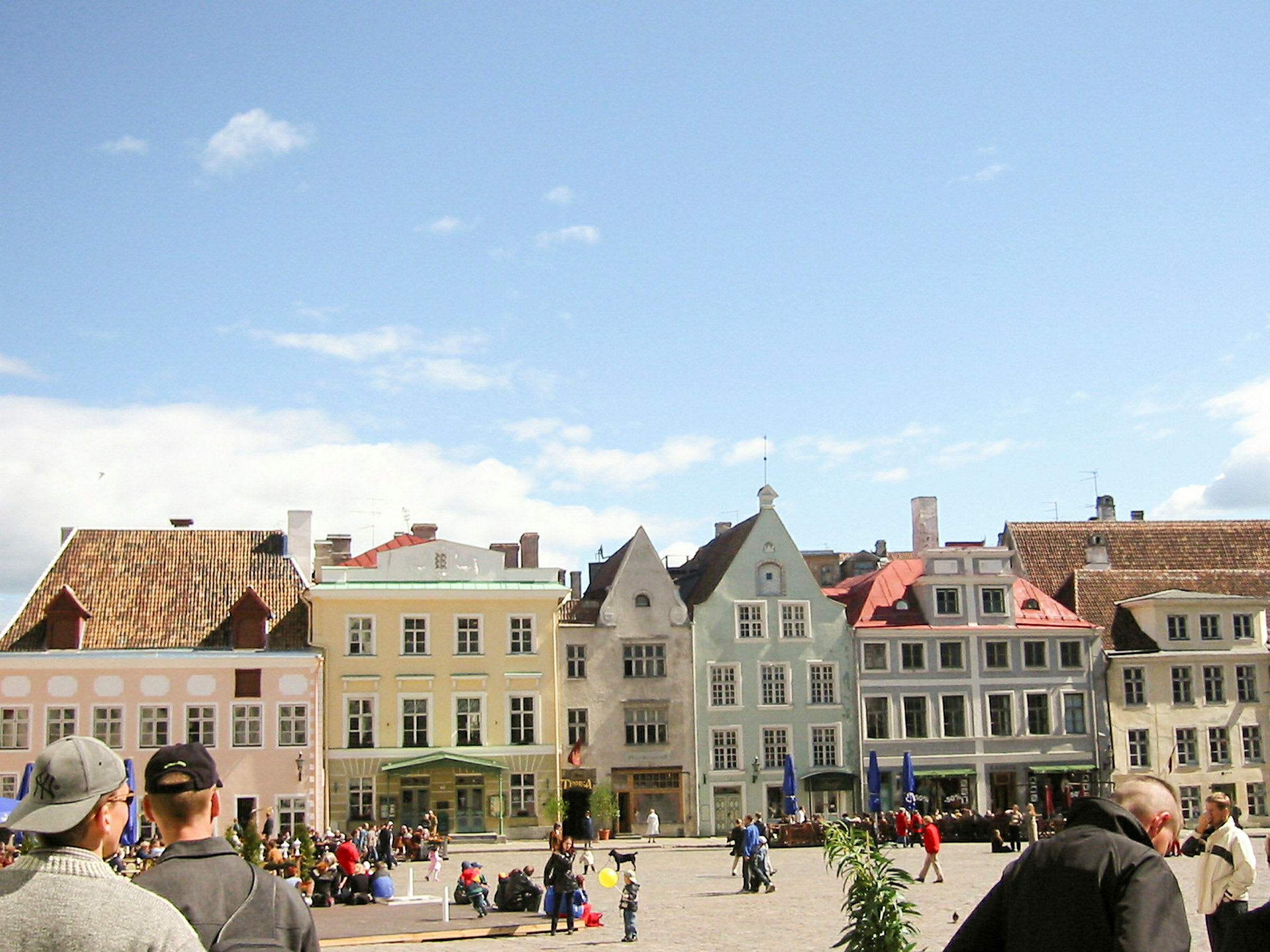 Et bytorg med fargerike bygninger i bakgrunnen og folk som spaserer og sitter på torget. (AI-generert tekst)
