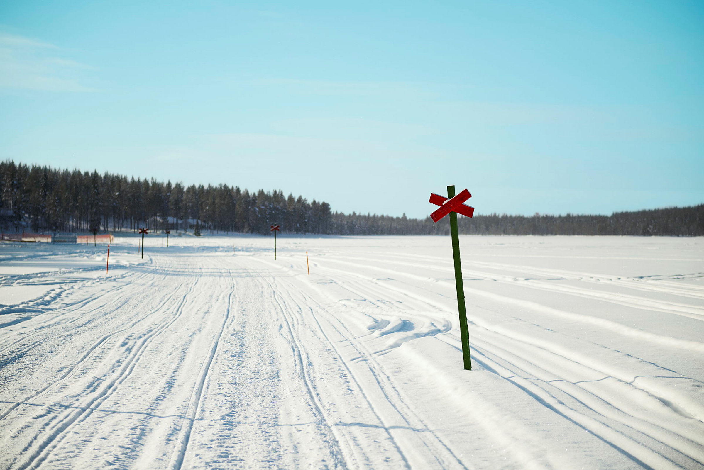En snødekt vei med røde kryssmerkede veiskilt og skog i bakgrunnen under en klar blå himmel. (AI-generert tekst)