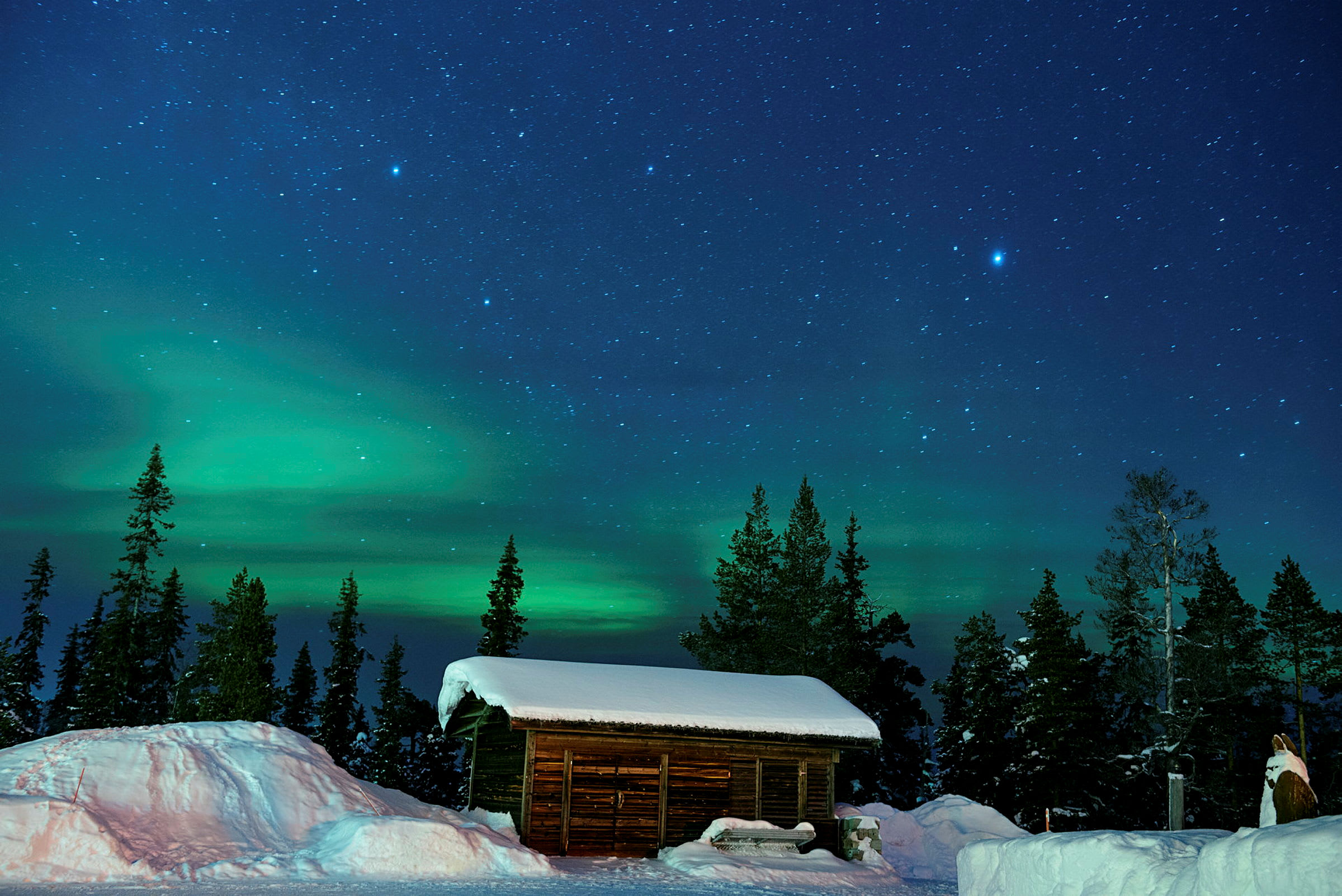 En tømmerhytte med snødekt tak foran en skog, med stjernehimmel og nordlys i bakgrunnen. (AI-generert tekst)