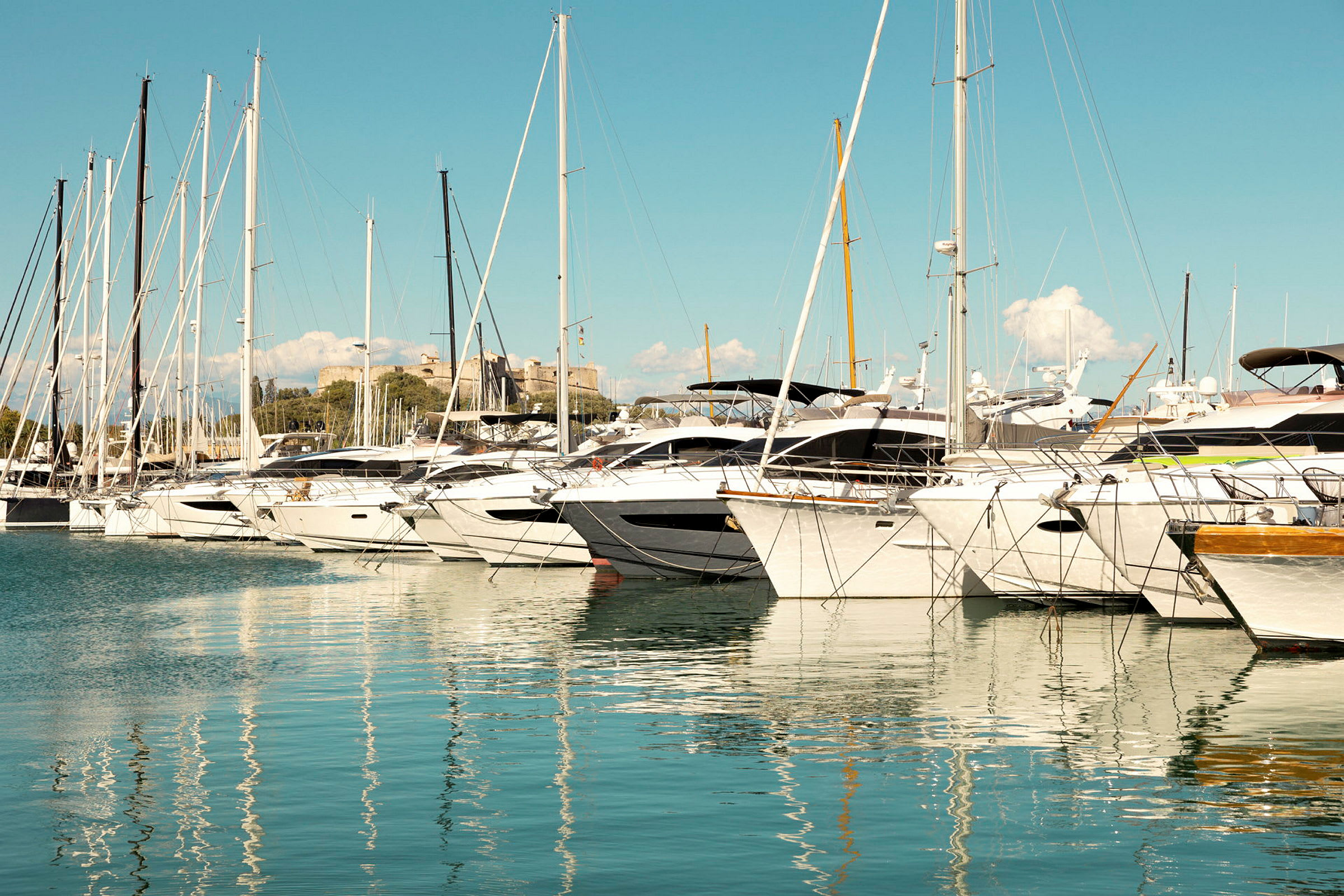 En rekke seilbåter ligger fortøyd i en marina. Vannet reflekterer båtene og mastene. (AI-generert tekst)