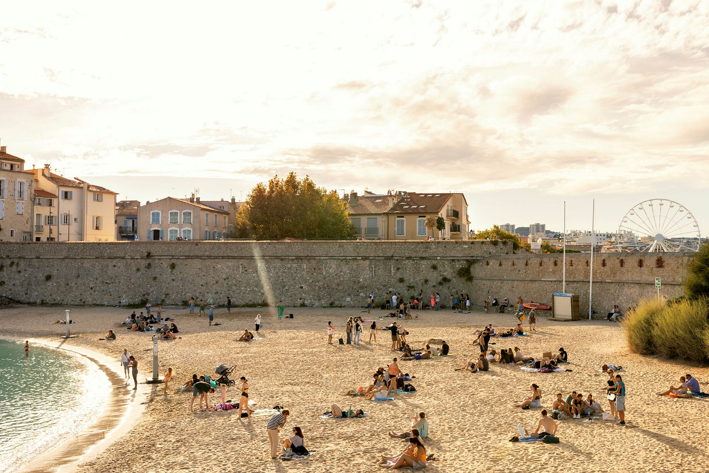 En strand med mennesker som slapper av på sanden, mens noen individer går langs vannkanten. Hus og en mur vises i bakgrunnen, samt et pariserhjul. (AI-generert tekst)