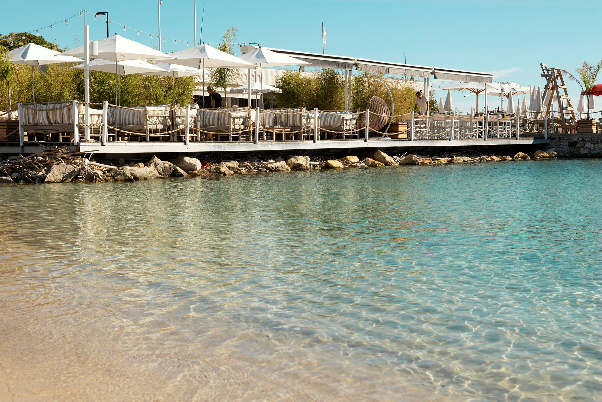 En restaurant med parasoller på en brygge ved vannet, med klart blått vann i forgrunnen. (AI-generert tekst)