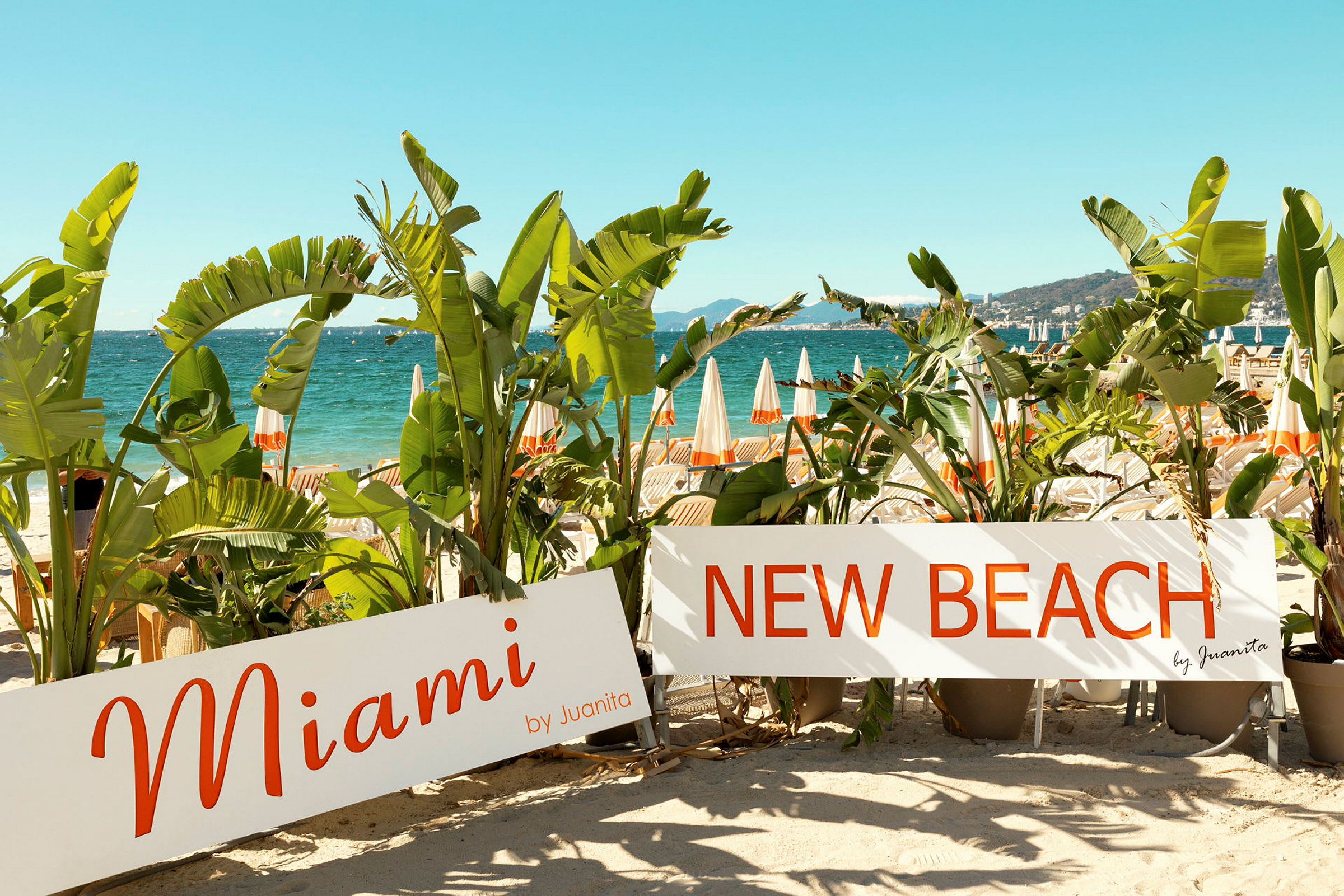 Stranden med parasoller i bakgrunnen, omgitt av palmer og skilt med tekstene "Miami" og "New Beach". (AI-generert tekst)