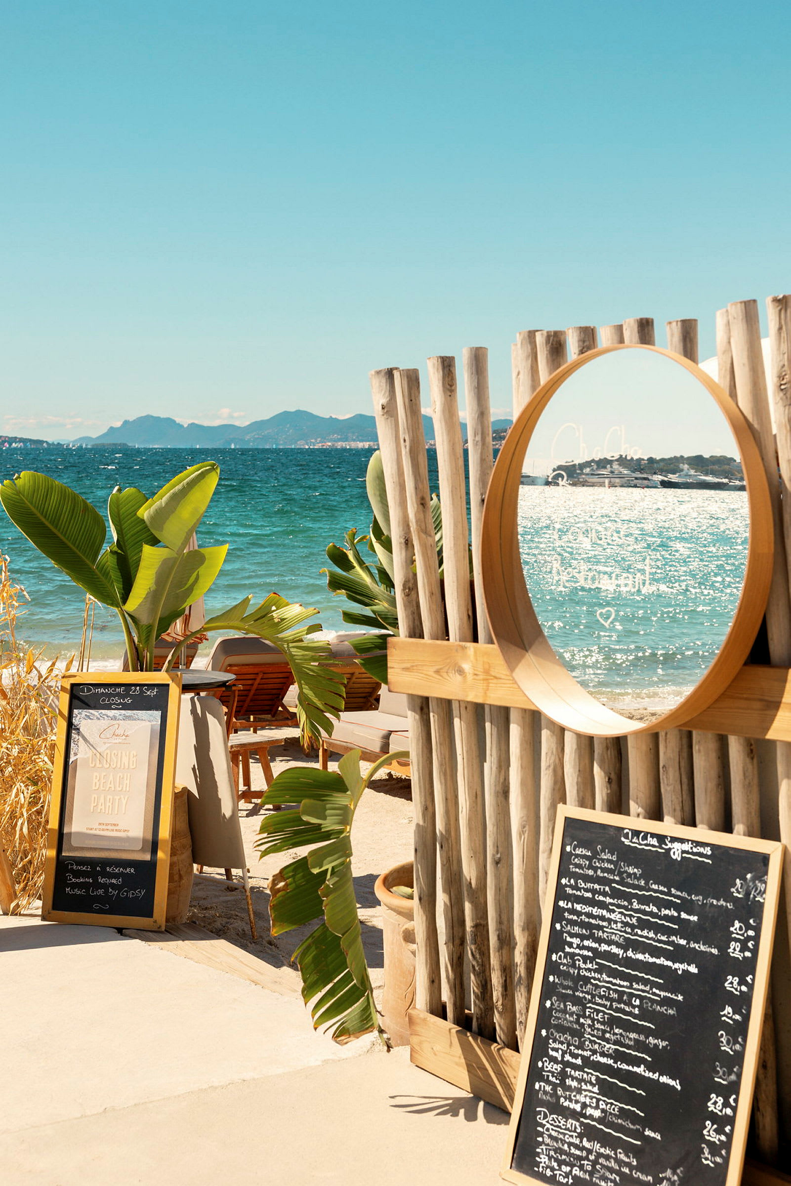 En strand med en treramme og et speil, omgitt av tropiske planter. En meny står på bakken ved siden av et skilt. (AI-generert tekst)