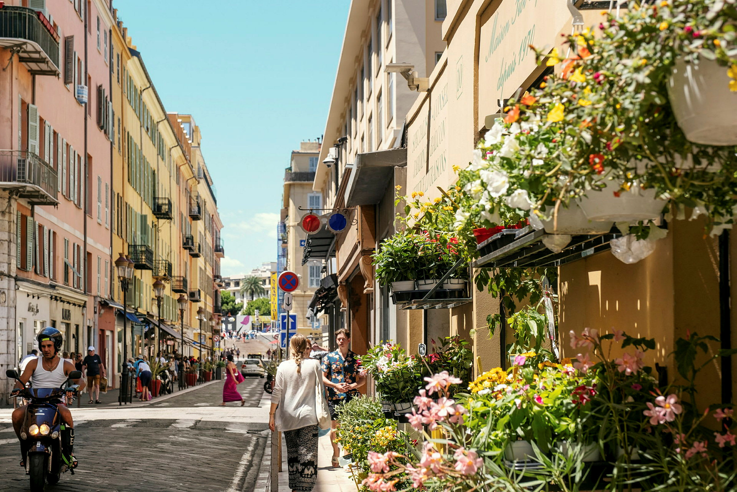 En bygate med stripete fargerike bygninger. Blomster henger på fasaden, og folk går og kjører moped på gaten. (AI-generert tekst)