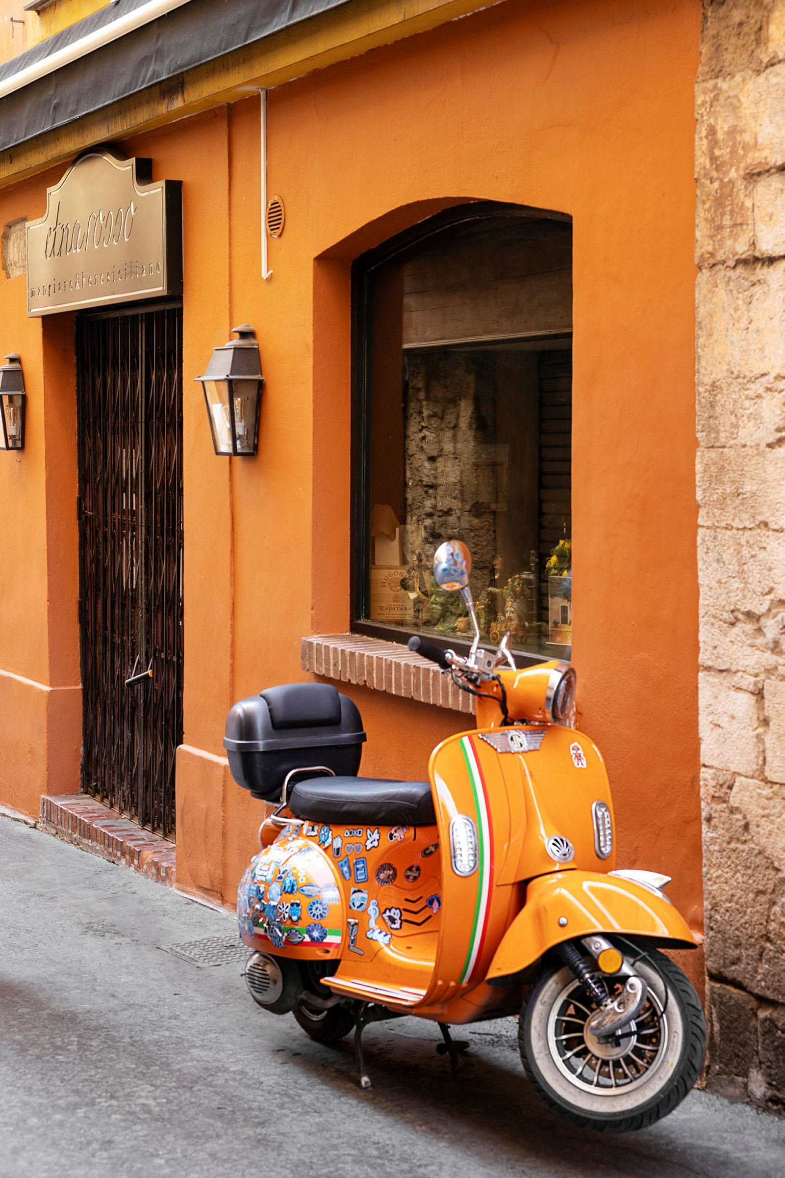 En oransje moped med dekorative klistremerker står parkert ved en oransje bygning med en smijernsport og et skiltet vindu. (AI-generert tekst)