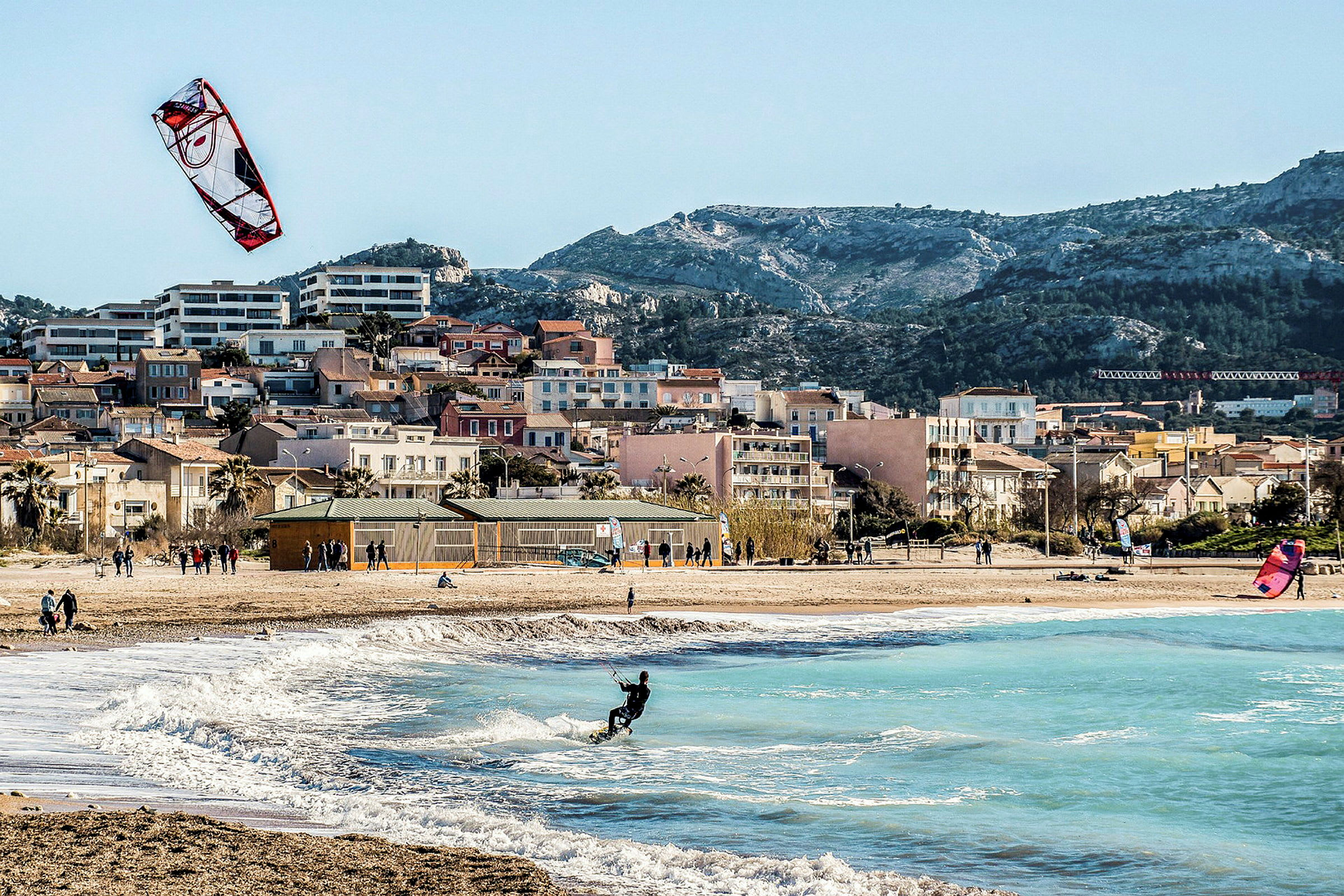 Strand med kitesurfere og bølger, hus synlige i bakgrunnen. (AI-generert tekst)