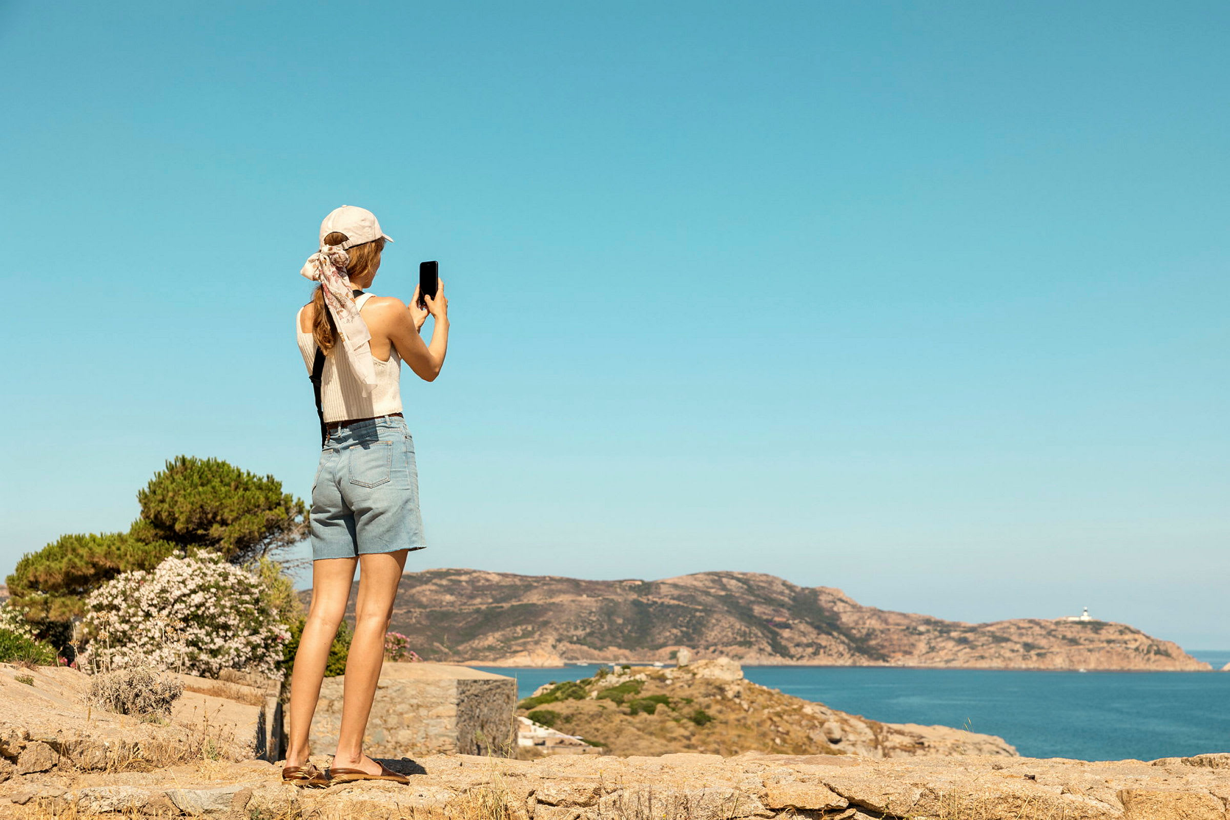 En person med hatt og shorts tar bilde av et kystlandskap med hav og klipper. (AI-generert tekst)