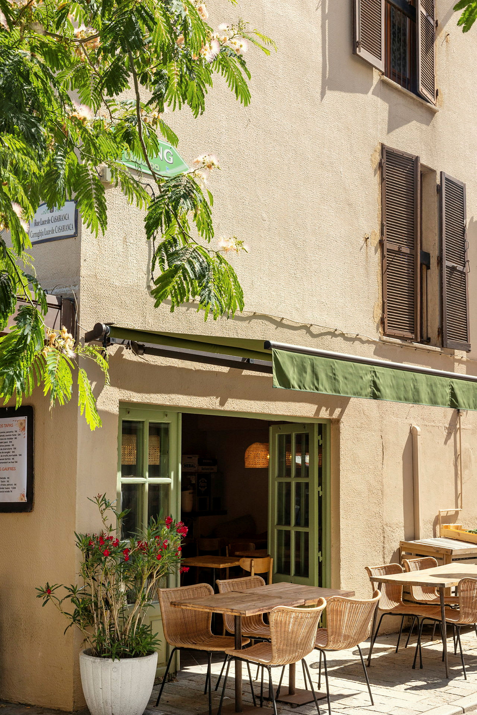 En kafé med uteservering, med bord og stoler plassert på en steinlagt gårdsplass. Grøntområder og blomster omgir bygningen. (AI-generert tekst)