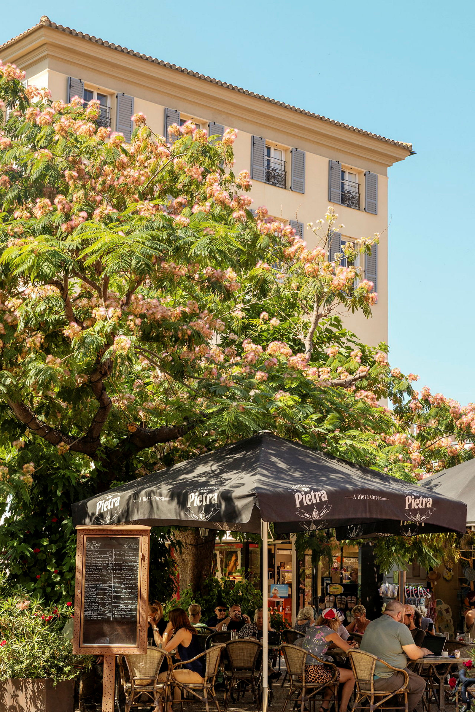 Bilde av et torg med servering under et tre med rosa blomster. Bygningen i bakgrunnen har blå skodder. (AI-generert tekst)