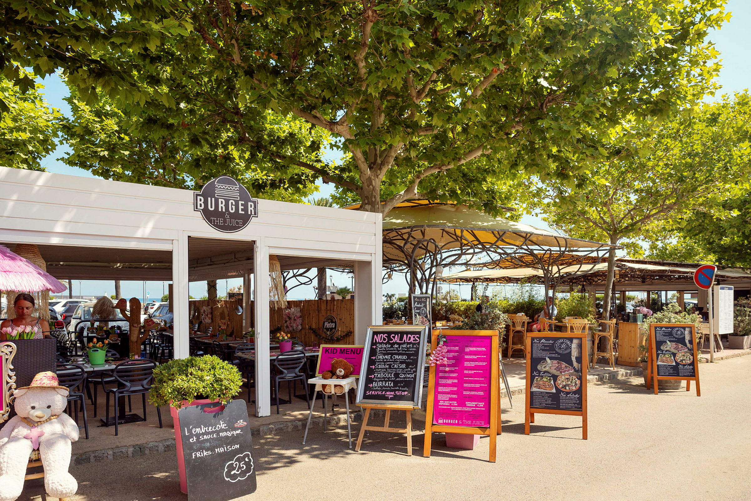 Utendørsrestaurant med skilt og beplantning. Bord og stoler står under trekroner og parasoller. (AI-generert tekst)