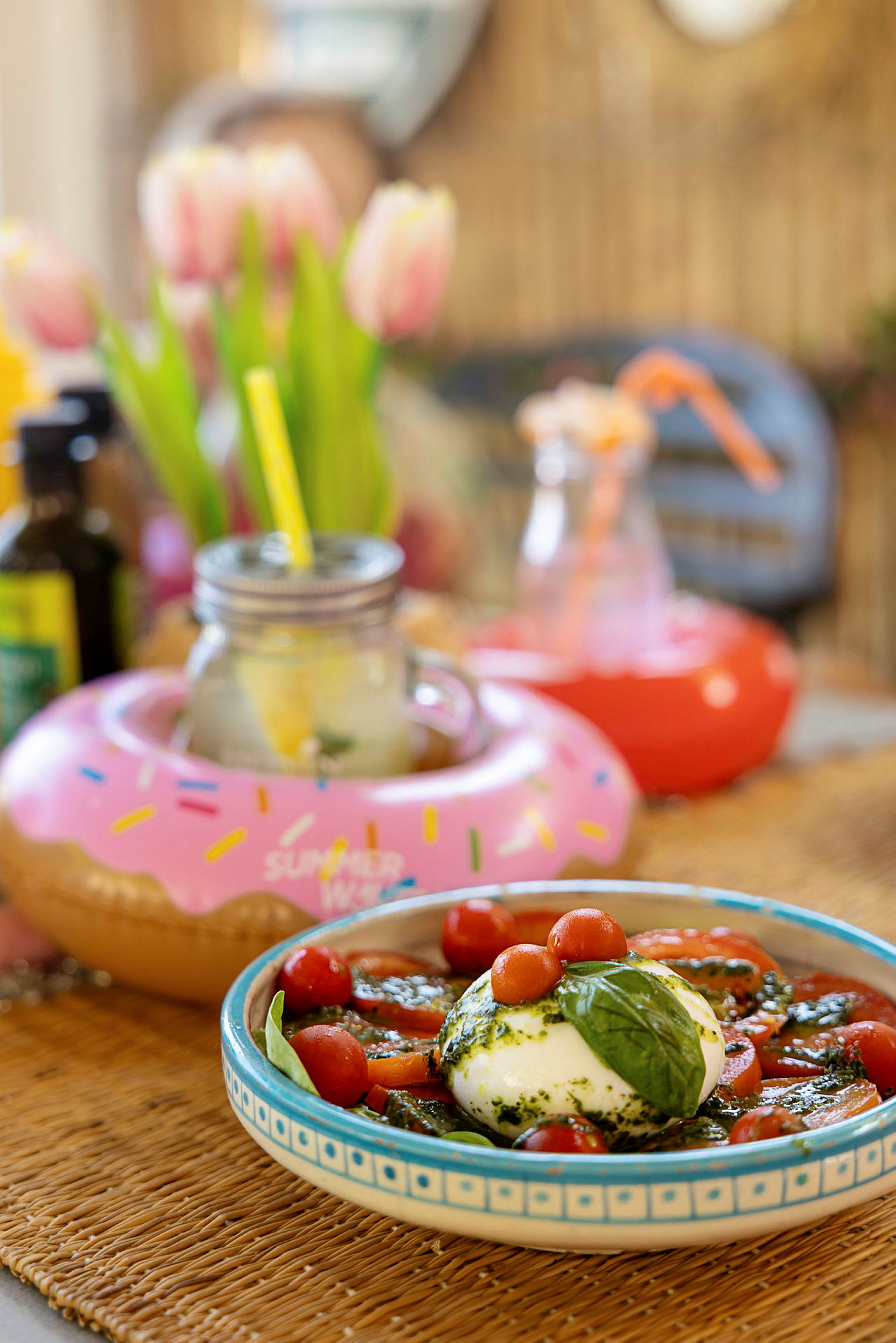 En rett med buffalo-mozzarella, tomater og basilikum samt drikker med dekorasjoner. Bakgrunnen viser tulipaner og andre dekorative gjenstander. (AI-generert tekst)