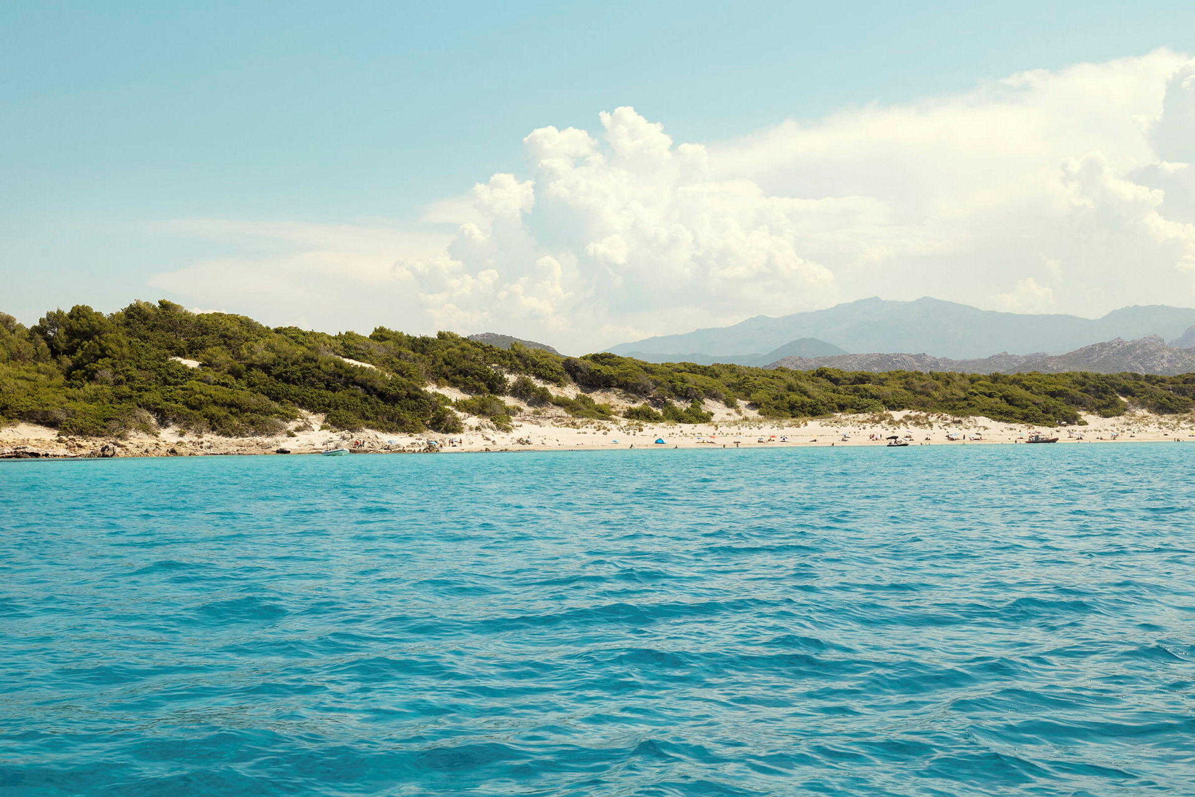 En strand med turkist vann, omgitt av grønn vegetasjon og fjell i bakgrunnen. (AI-generert tekst)