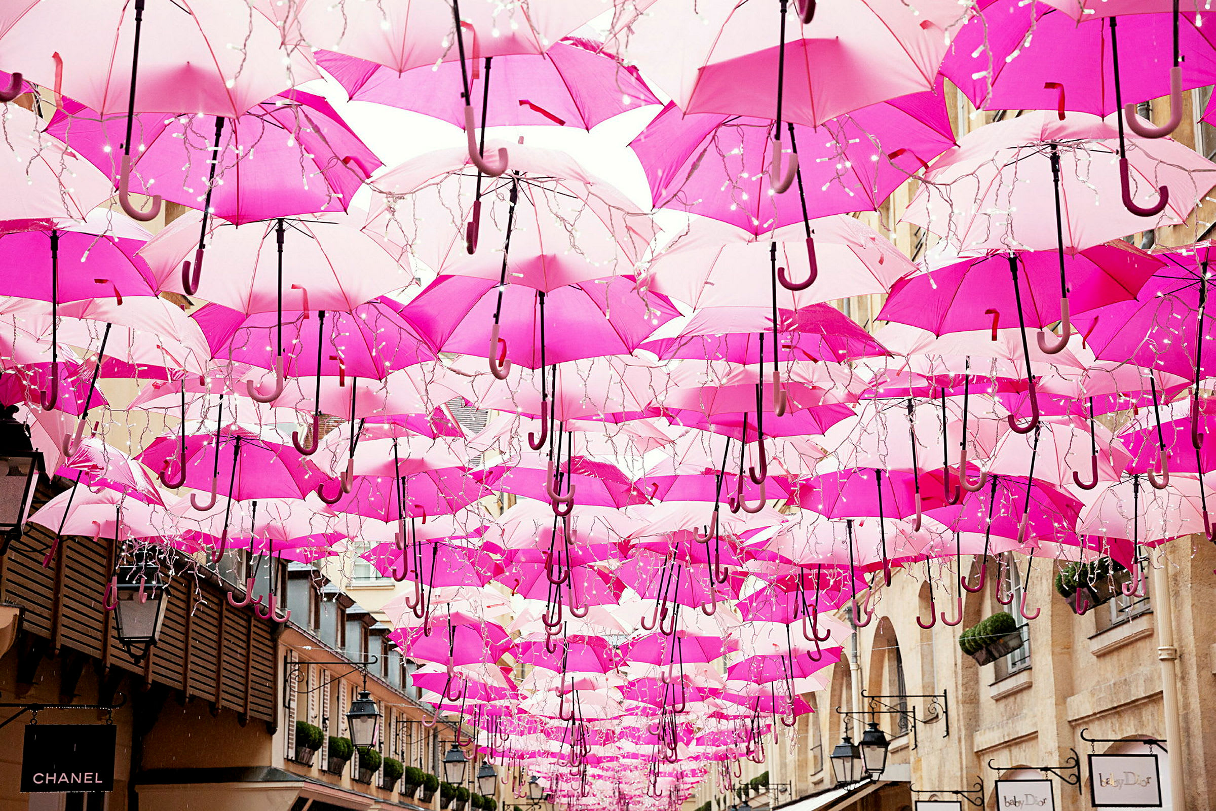 Flere rosa paraplyer henger opp i luften mellom bygninger. (AI-generert tekst)