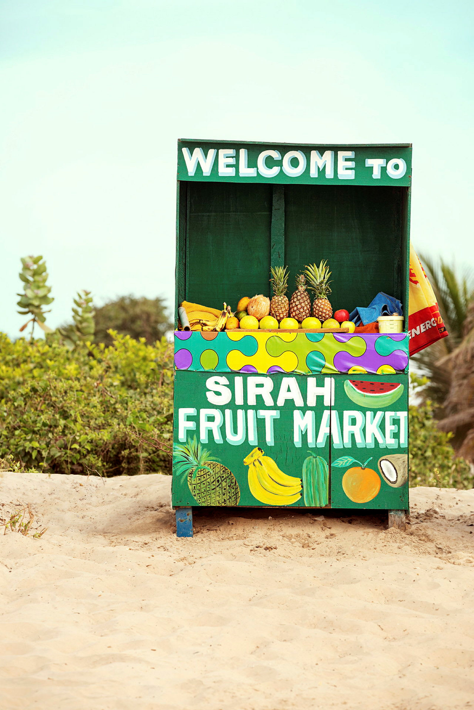 En fruktbod med teksten "Sirah Fruit Market" tilbyr en rekke frukter, inkludert ananas, bananer og sitrusfrukter. Boden står på sand med vegetasjon i bakgrunnen. (AI-generert tekst)