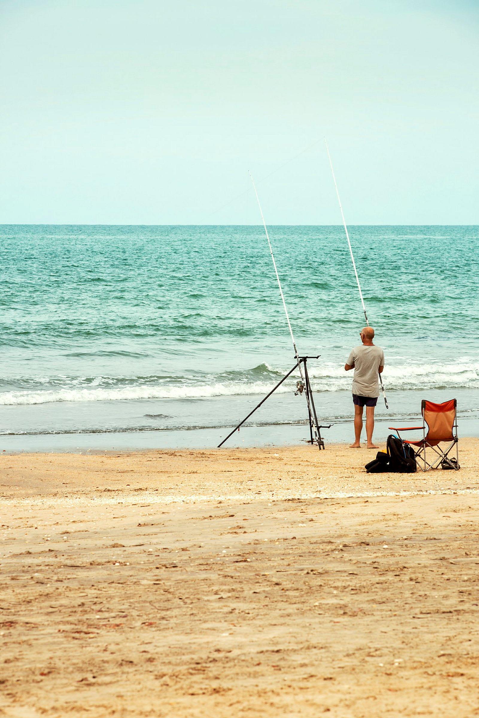 En person står ved strandkanten med to fiskestenger på et stativ. En stol står i sanden ved siden av. (AI-generert tekst)