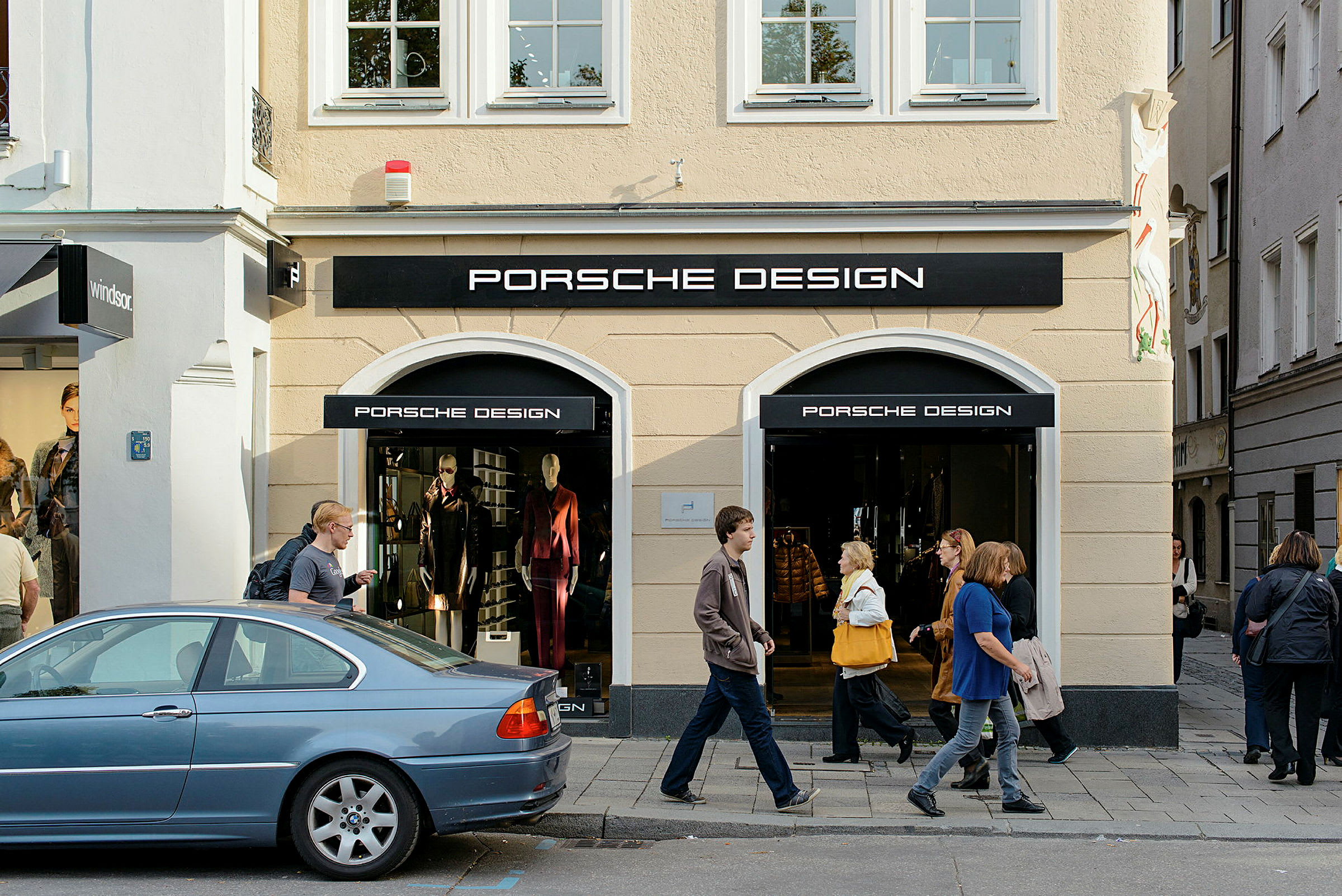 Inngang til en butikk med Porsche Design-logo, med utstillingsvinduer og folk som går på fortauet utenfor. (AI-generert tekst)
