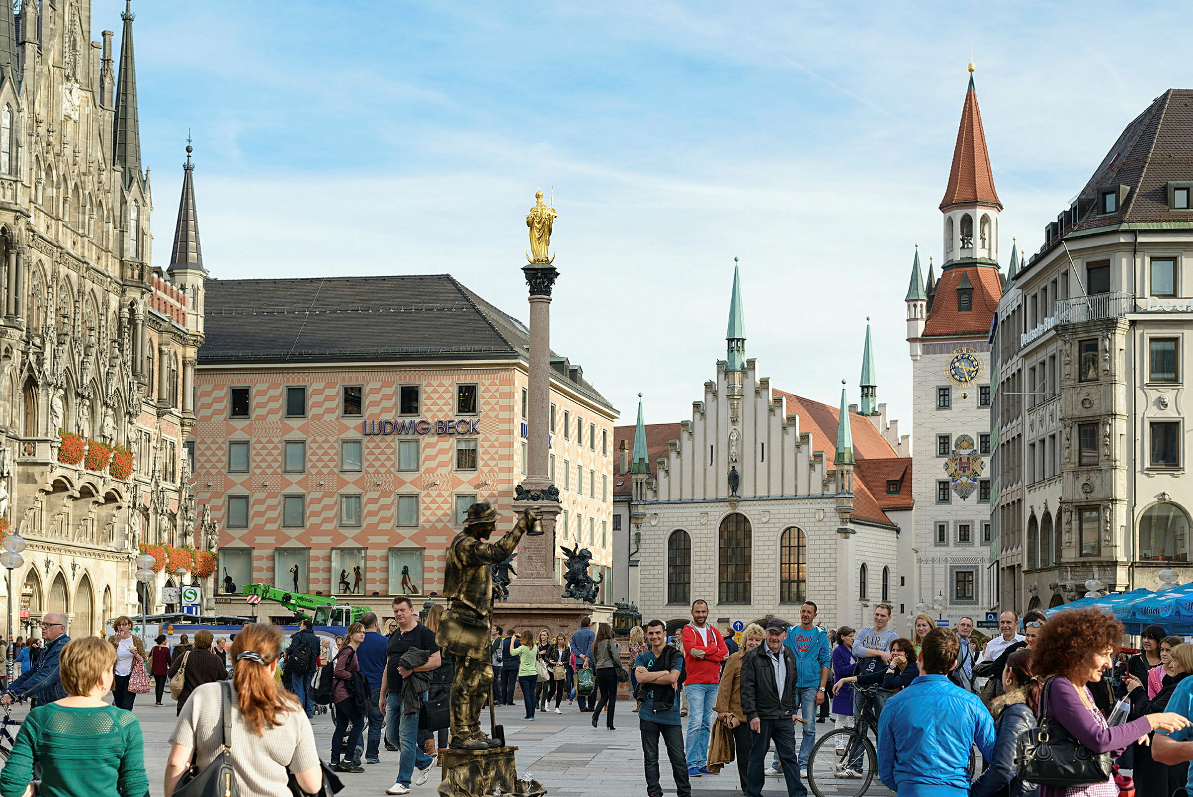 Et livlig torg med historiske bygninger, en statue med en gyllen topp og mange gående personer. (AI-generert tekst)