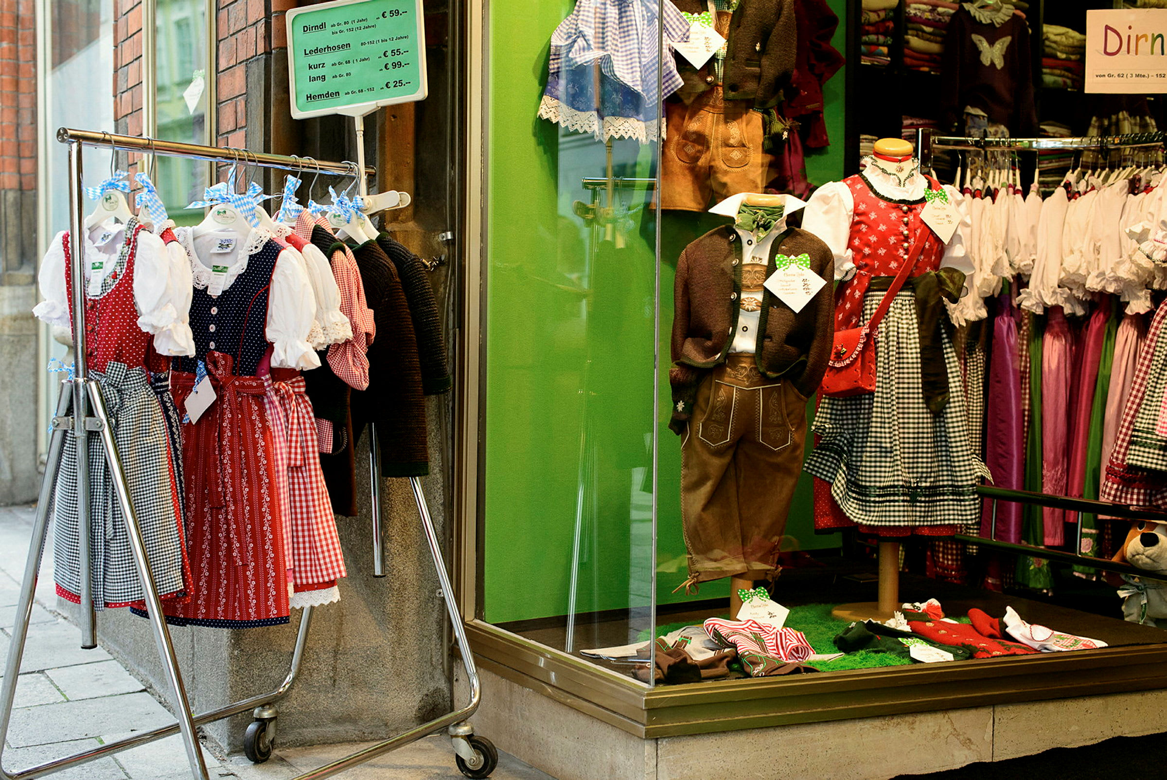 Et butikkvindu med tradisjonelle folkedrakter og et klesstativ utenfor fylt med dirndl-kjoler. (AI-generert tekst)