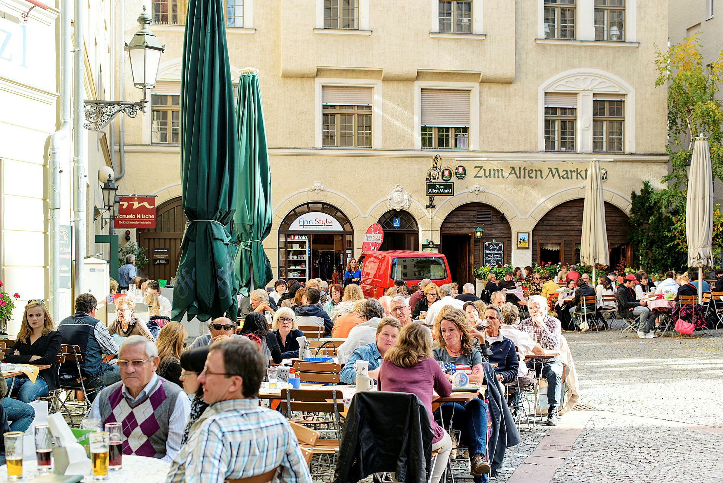 Utendørs servering med mange mennesker sittende ved trebord på en brosteinsbelagt patio, omgitt av gamle bygninger. (AI-generert tekst)