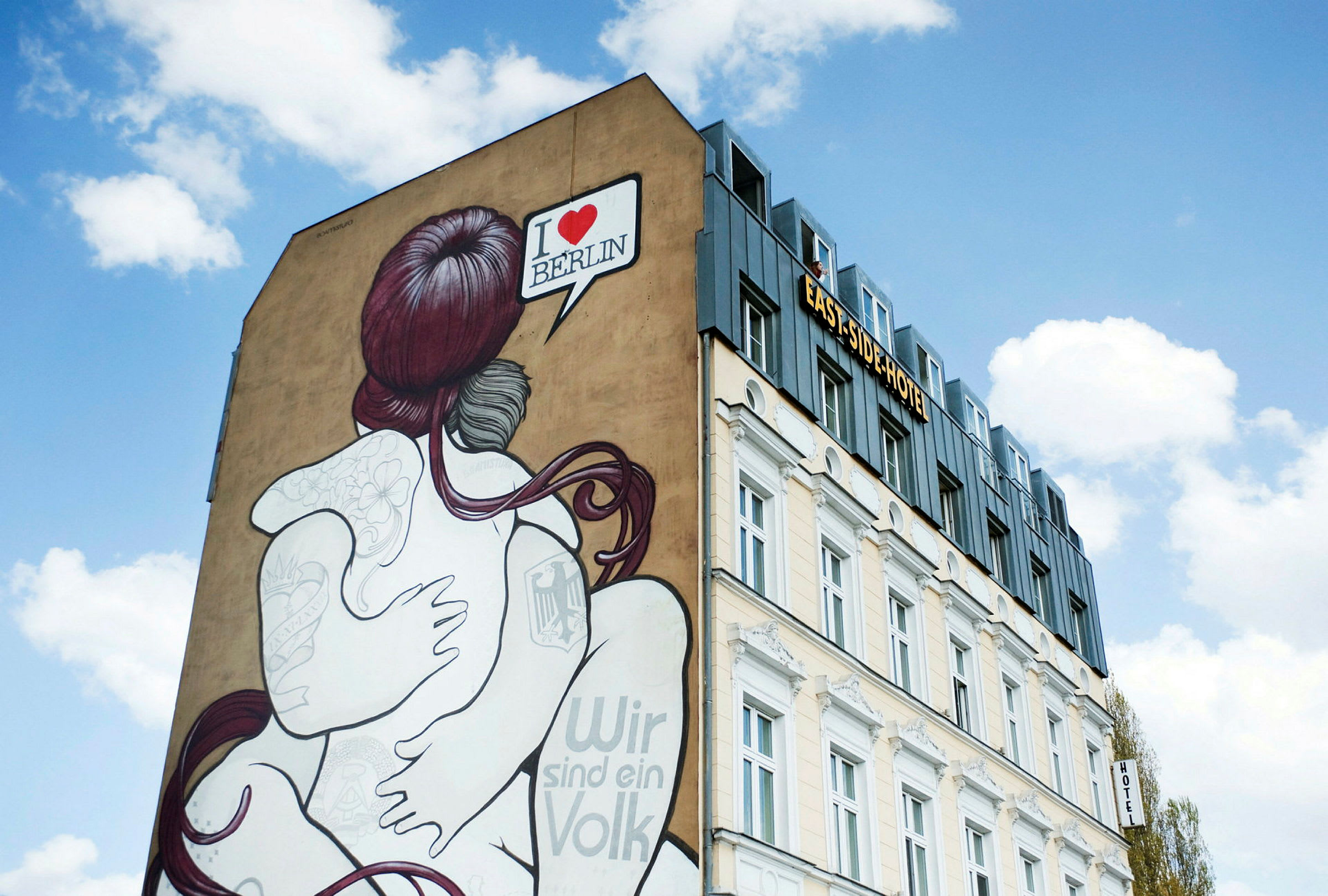 En bygning med et veggmaleri av et par som omfavner hverandre. Teksten "I ♥ BERLIN" vises i en snakkeboble. Himmlen er klar med hvite skyer. (AI-generert tekst)