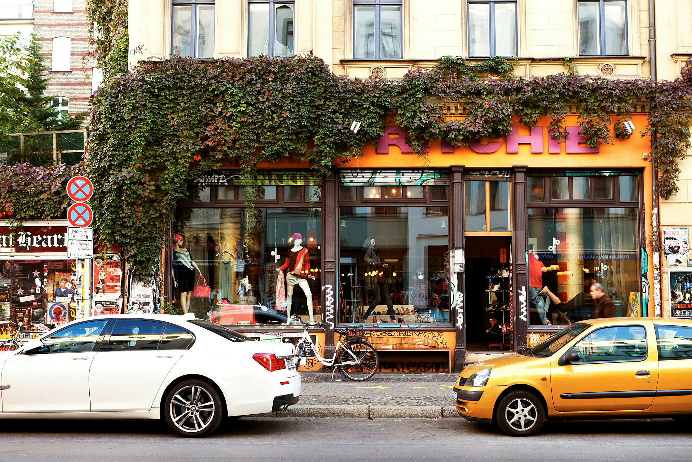 En bygning med en butikk har et oransje skilt med store bokstaver, omgitt av grønne planter som klatrer langs fasaden. Biler står parkert foran butikken. (AI-generert tekst)
