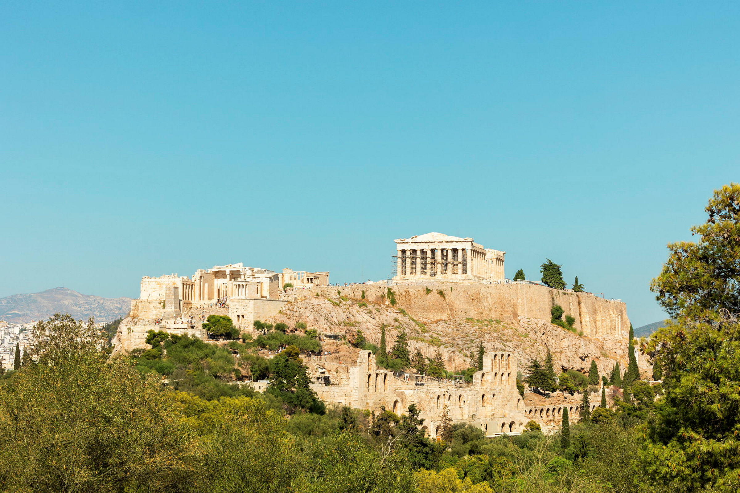 Fotografi av Akropolis med Parthenon på toppen, omgitt av grøntområder og med klar blå himmel i bakgrunnen. (AI-generert tekst)