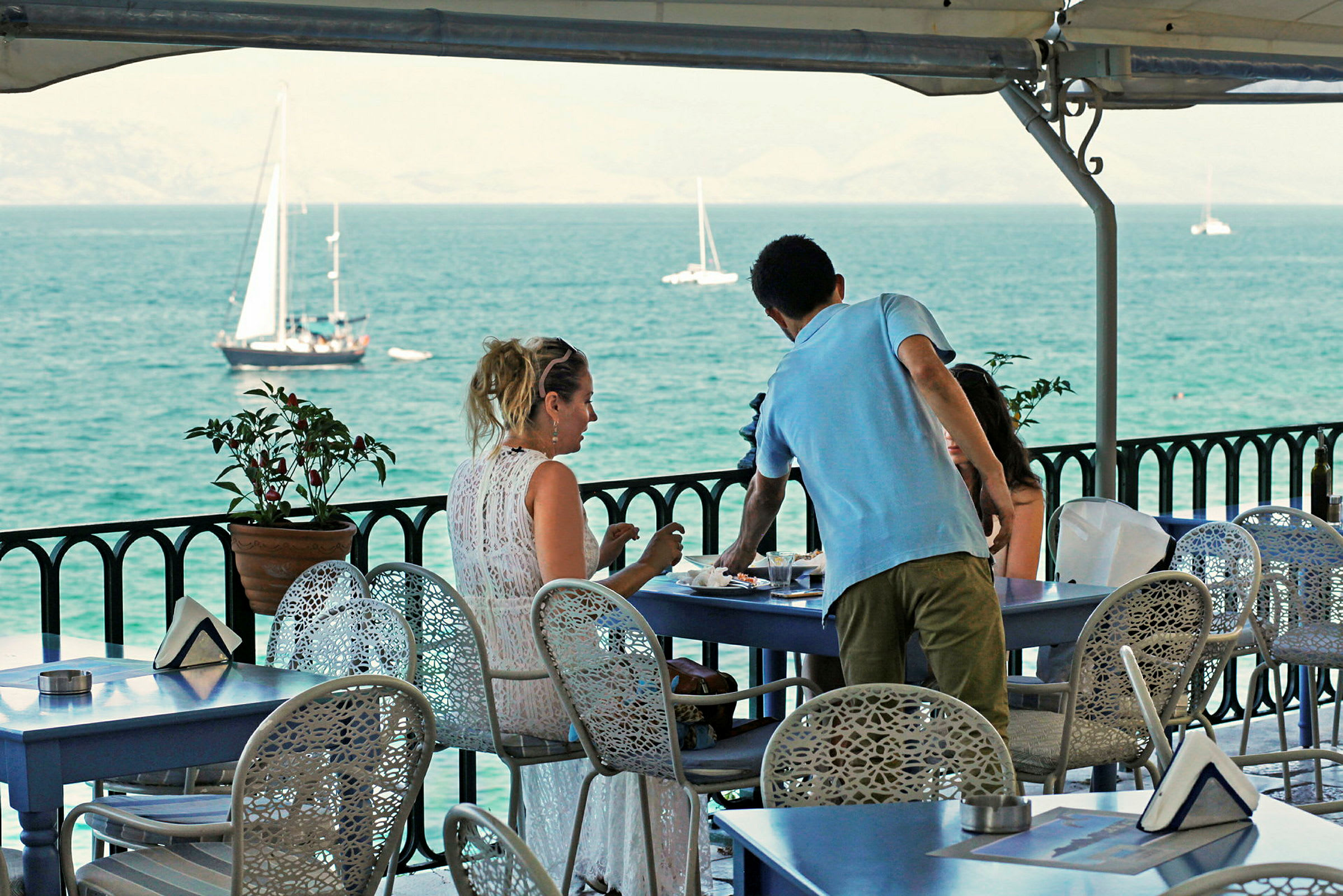 Folk sitter ved et bord på en terrasse med utsikt over havet og seilbåter. (AI-generert tekst)