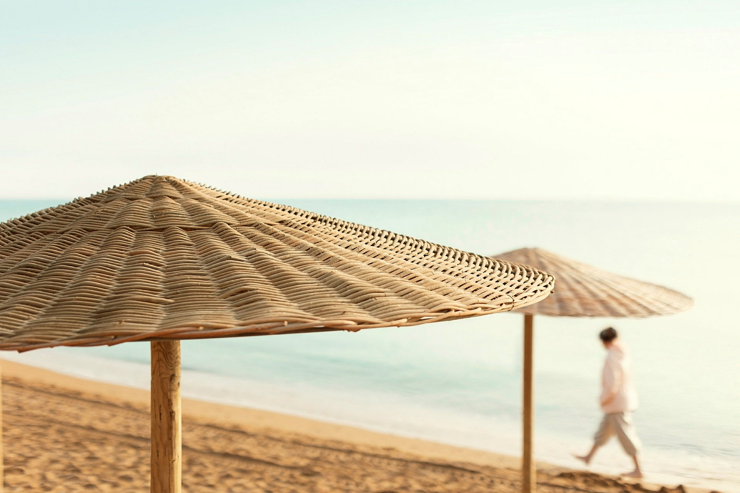 Strand med to solparasoller av rotting og en person som går langs vannet. (AI-generert tekst)