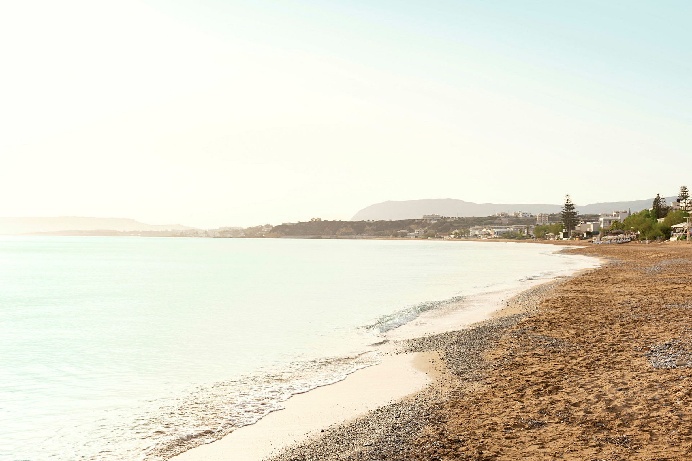 Strand med sand og småstein, med havet ved siden og bygninger i bakgrunnen. (AI-generert tekst)