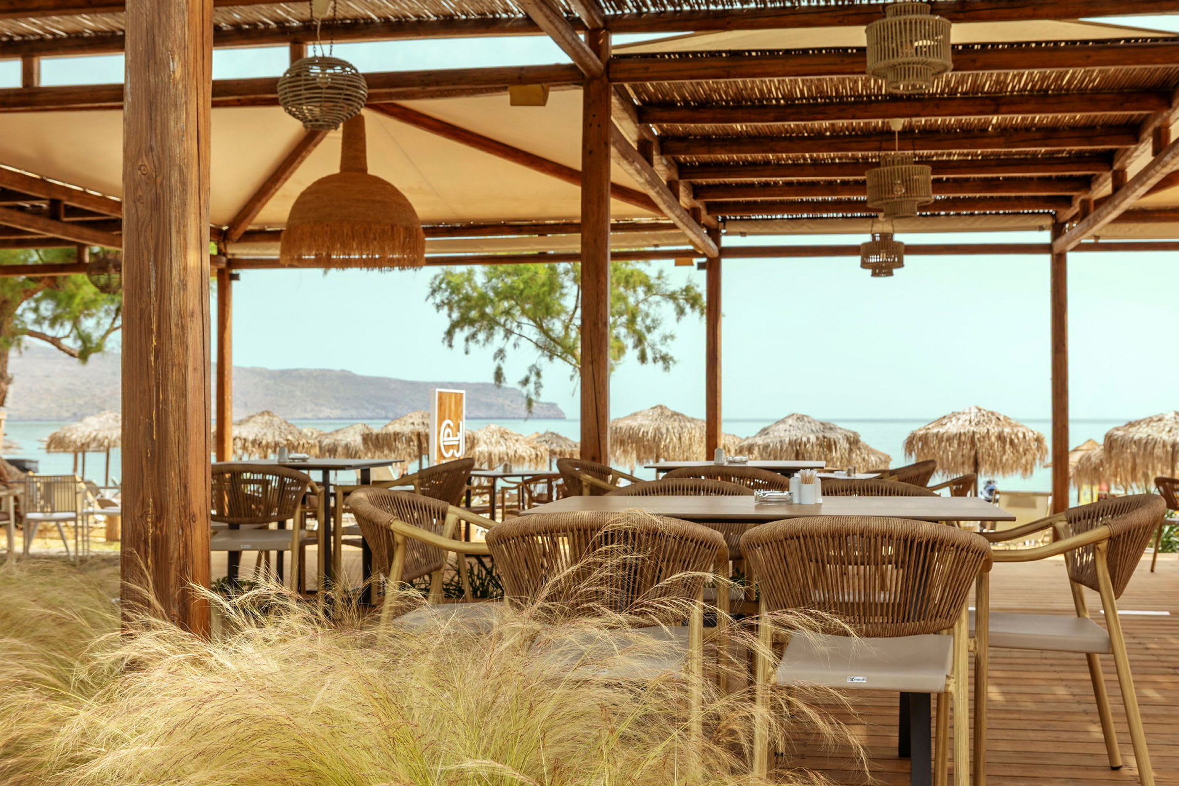 En utendørs restaurant med bord og stoler under en treskjerm. Bakgrunnen viser stranden og havet. (AI-generert tekst)