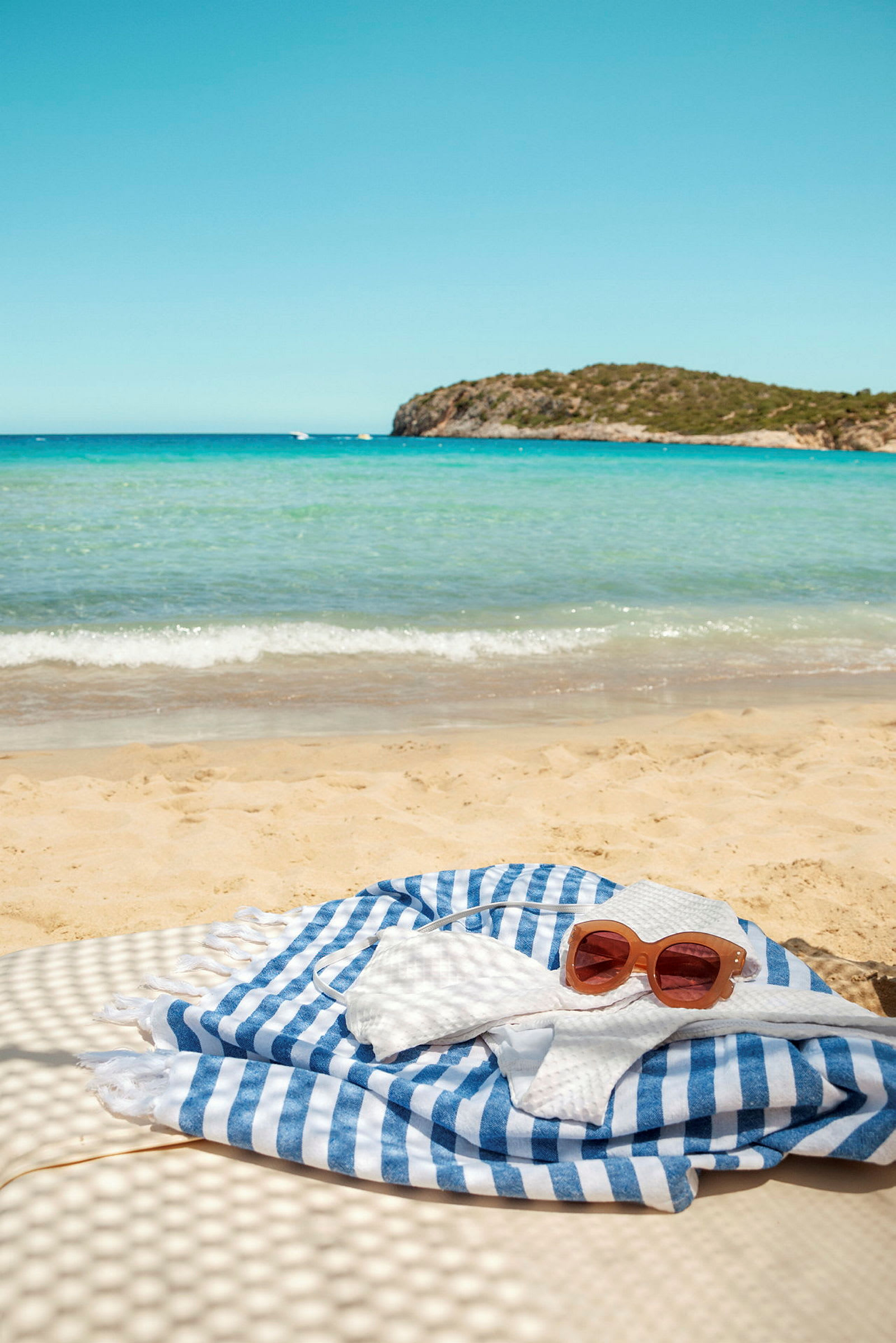Sandstrand ved havet med klipper i bakgrunnen og stripet håndkle i forgrunnen. (AI-generert tekst)