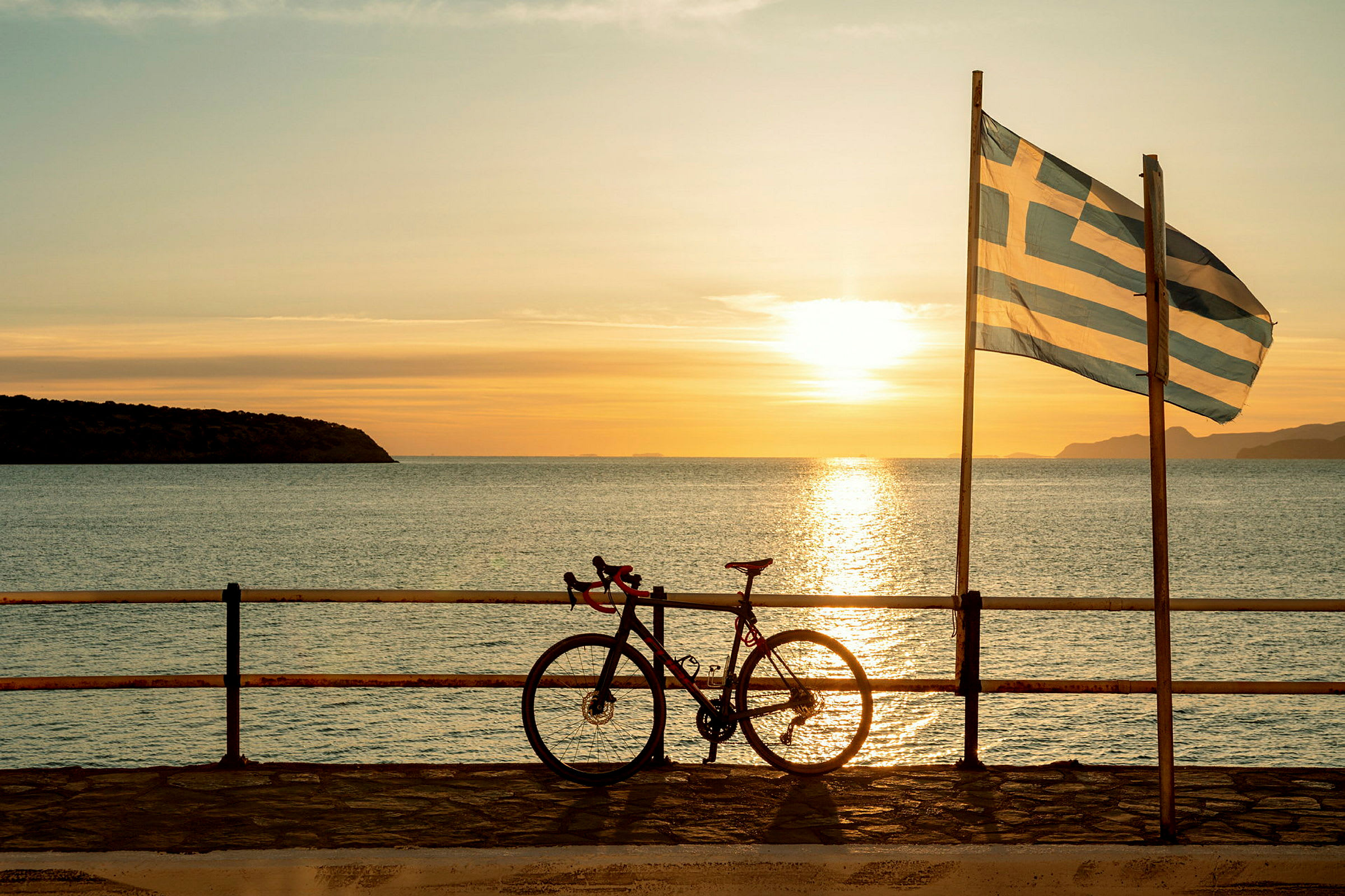 En sykkel står lent mot et rekkverk ved havet med et gresk flagg og en solnedgang i bakgrunnen. (AI-generert tekst)