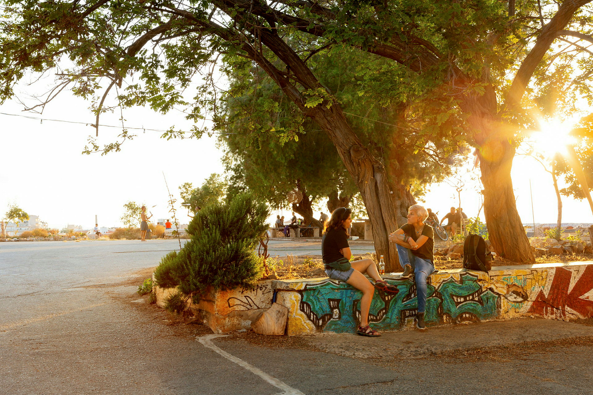 Personer sitter på en graffiti-dekorert mur under et tre, med solnedgang i bakgrunnen. (AI-generert tekst)