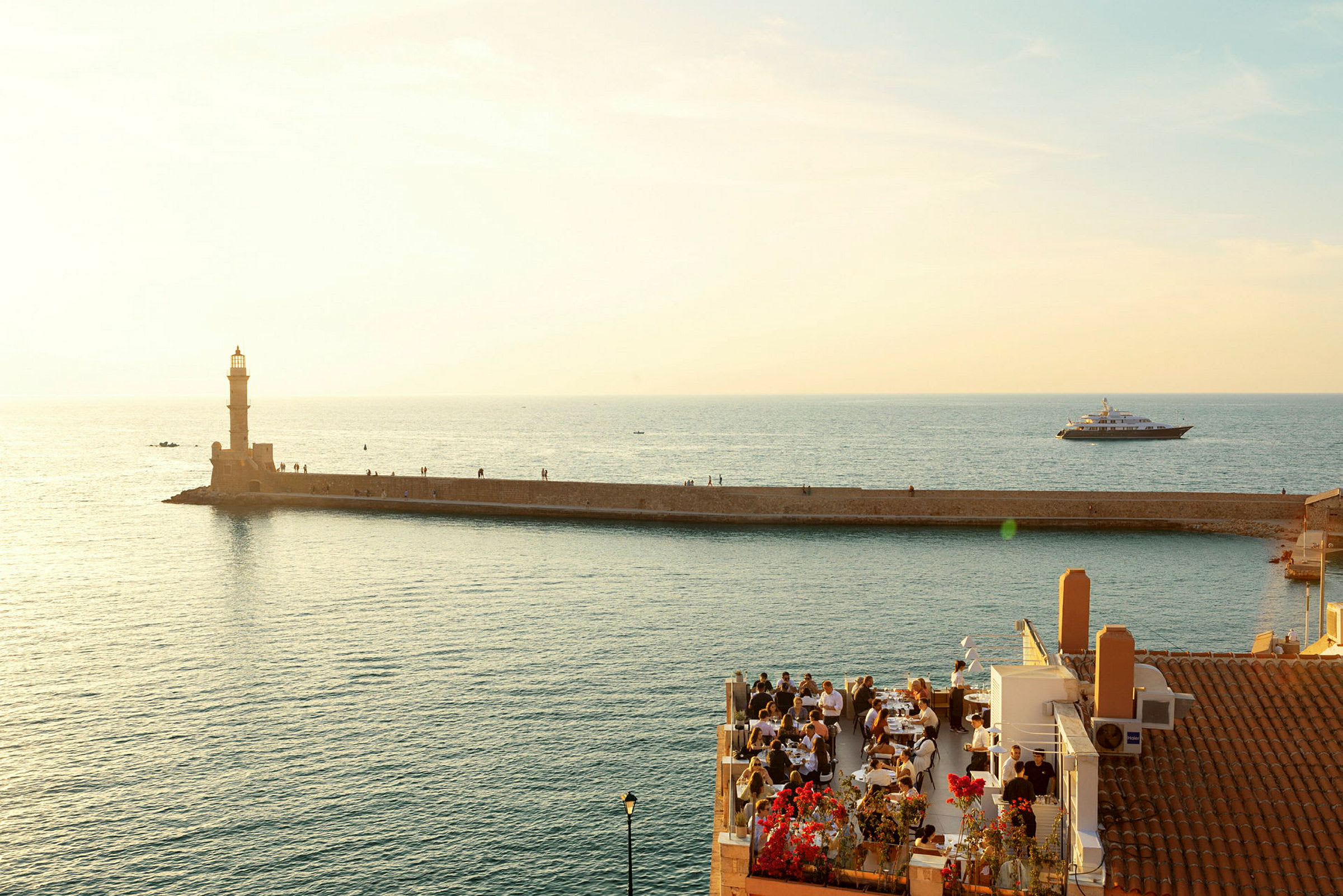 Et fyrtårn på en molo strekker seg ut mot havet i et solopplyst område med restaurantgjester i forgrunnen. (AI-generert tekst)