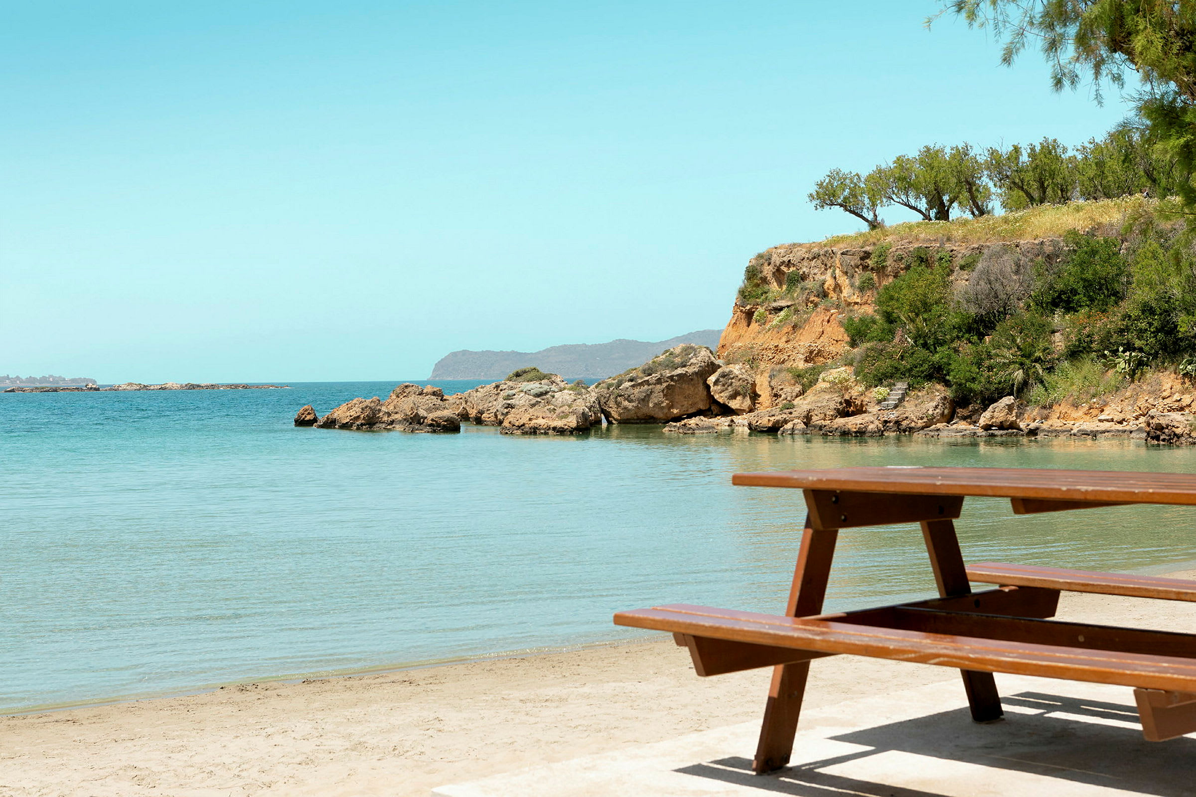 Piknikbord ved en sandstrand med klart vann og klippeformasjoner i bakgrunnen. (AI-generert tekst)