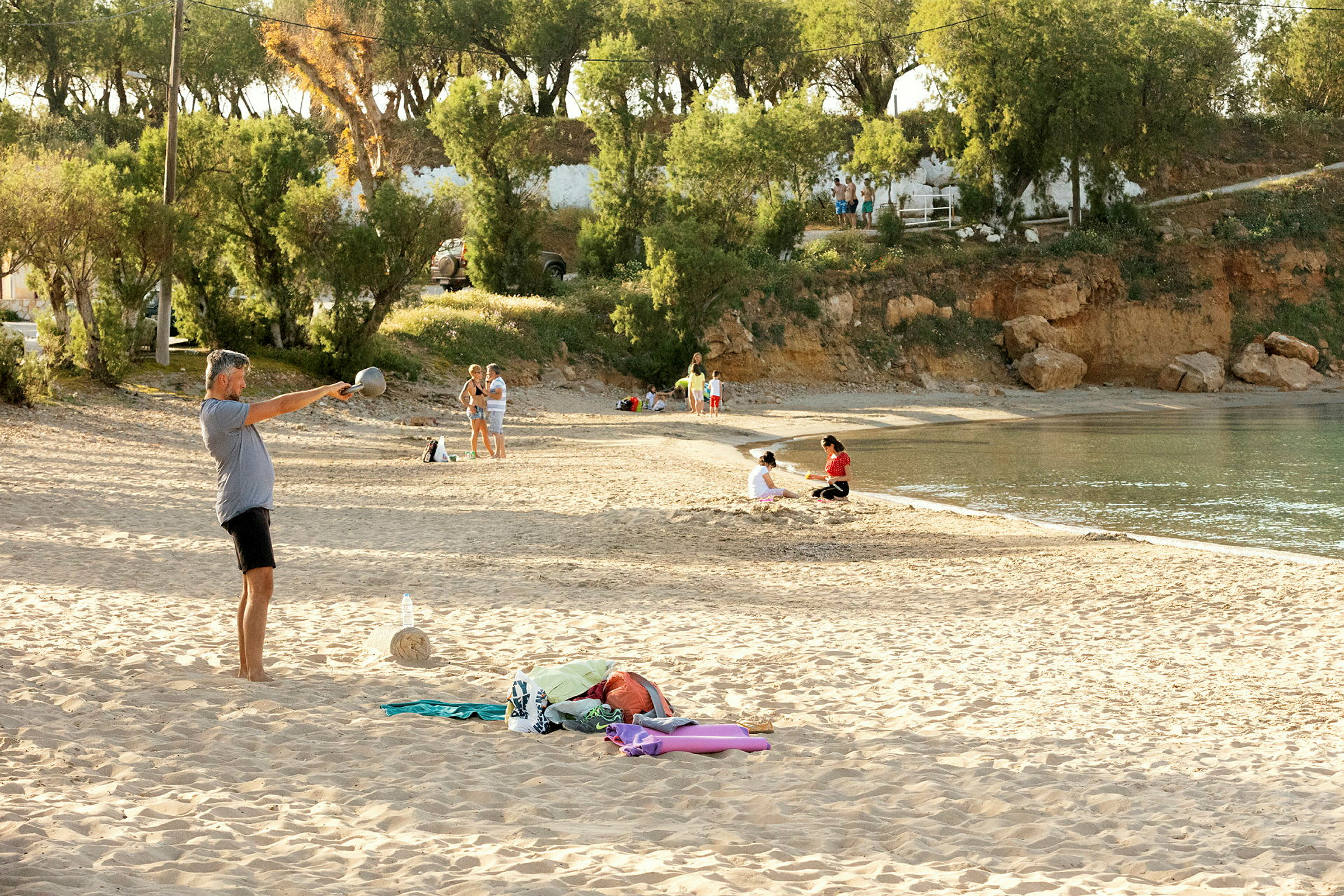 Strand med mennesker på sanden og ved vannkanten, omgitt av trær og klipper. (AI-generert tekst)