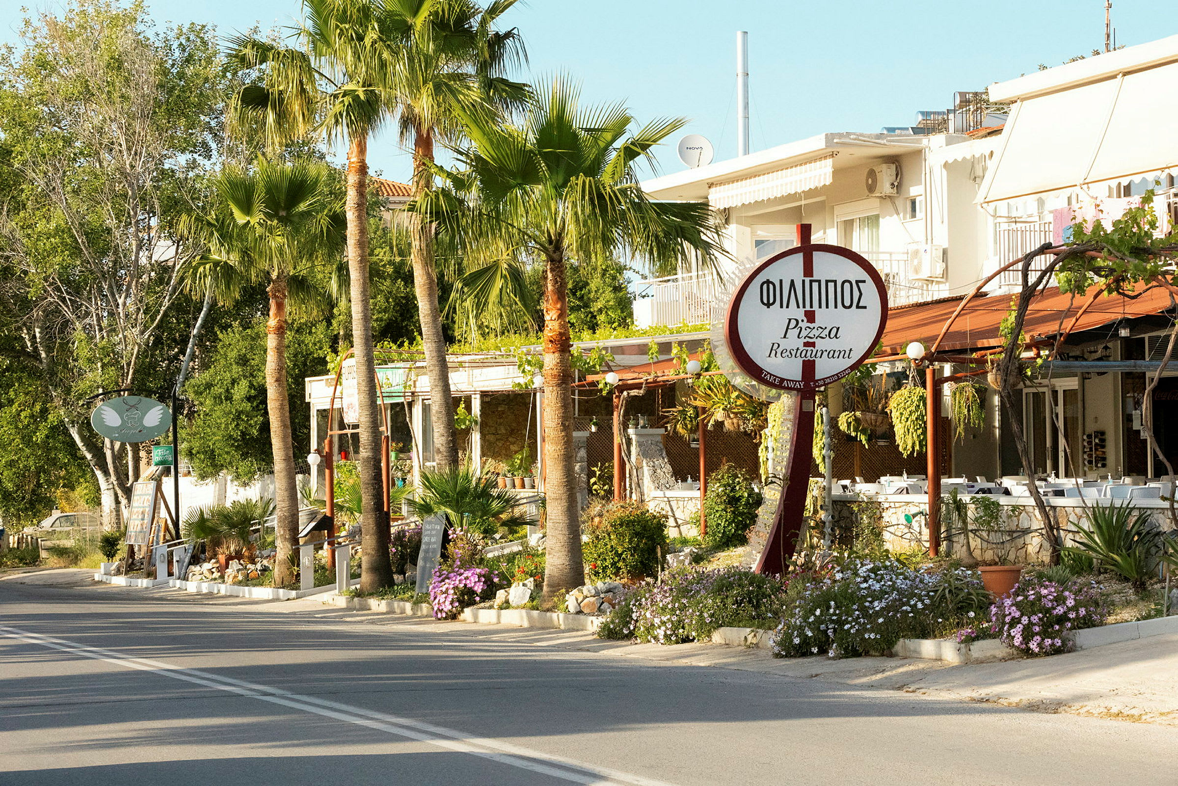 En vei kantet med palmer passerer forbi en restaurant med et skilt som viser navnet 'Philippos Pizza Restaurant'. (AI-generert tekst)