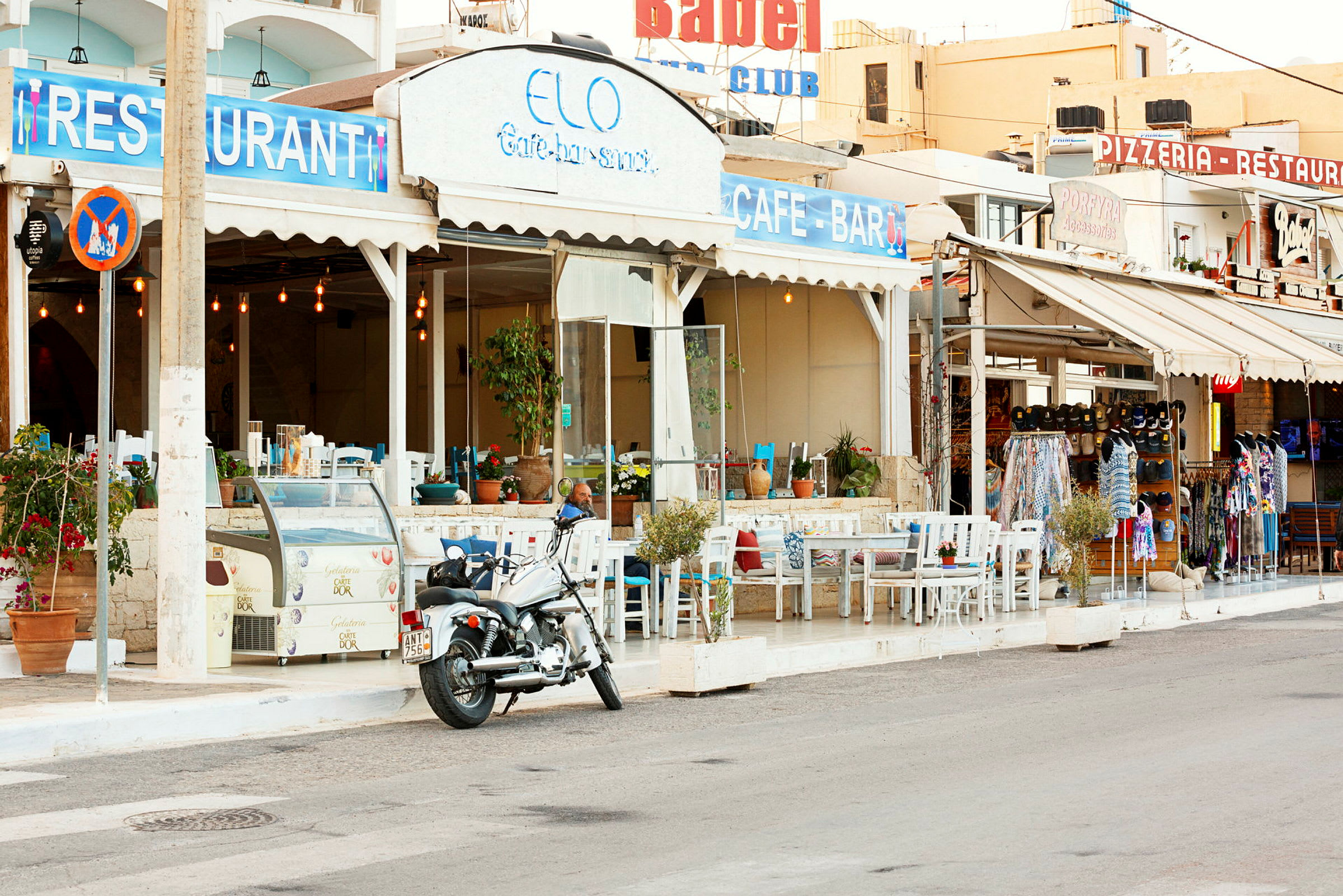 Motiv av en kafé med uteservering ved en gate, et skilt med teksten "Restaurant" og en motorsykkel parkert i forgrunnen. (AI-generert tekst)